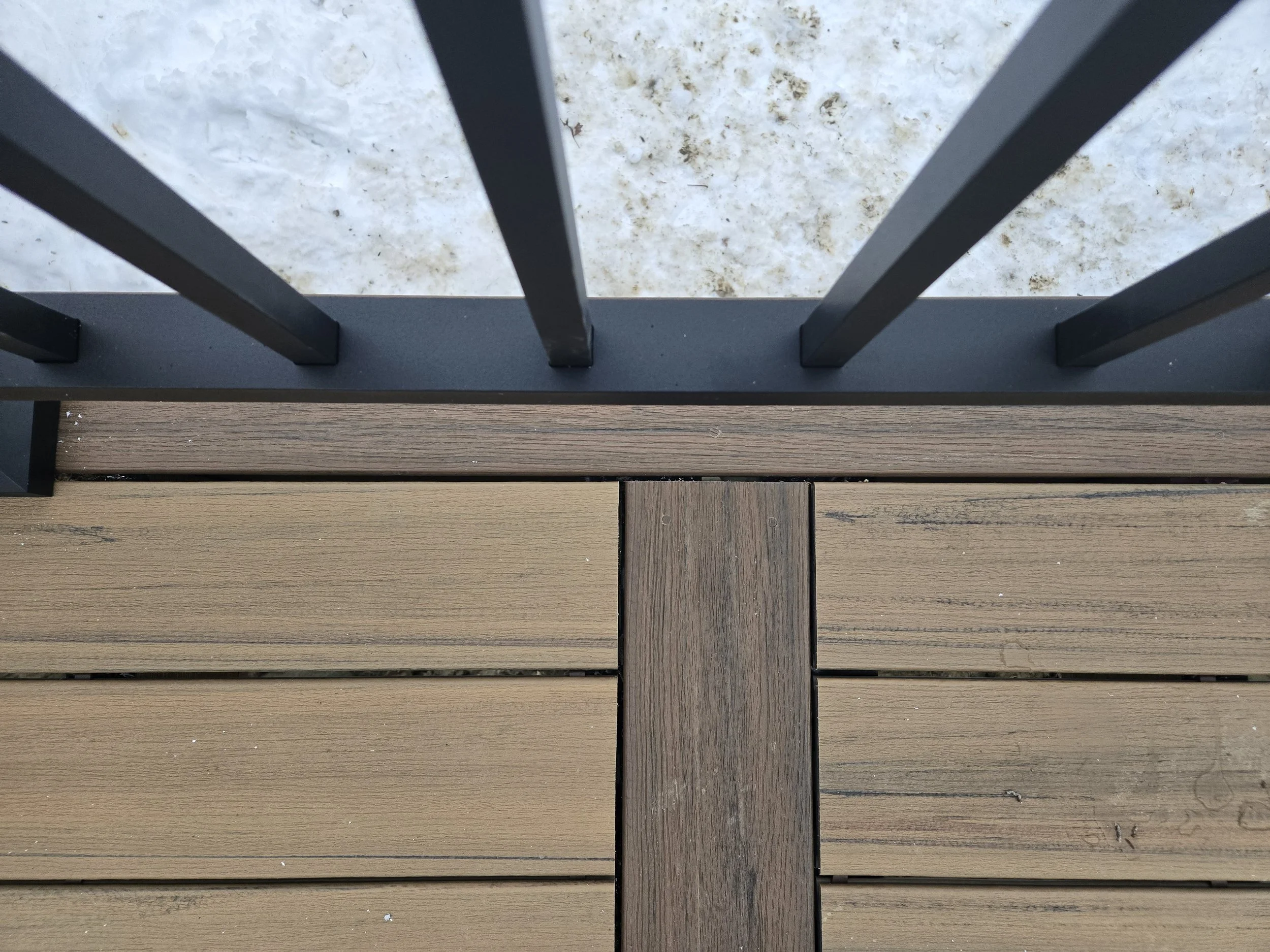 Close-up of a brown wooden deck with black metal railing and snow on the ground beyond.
