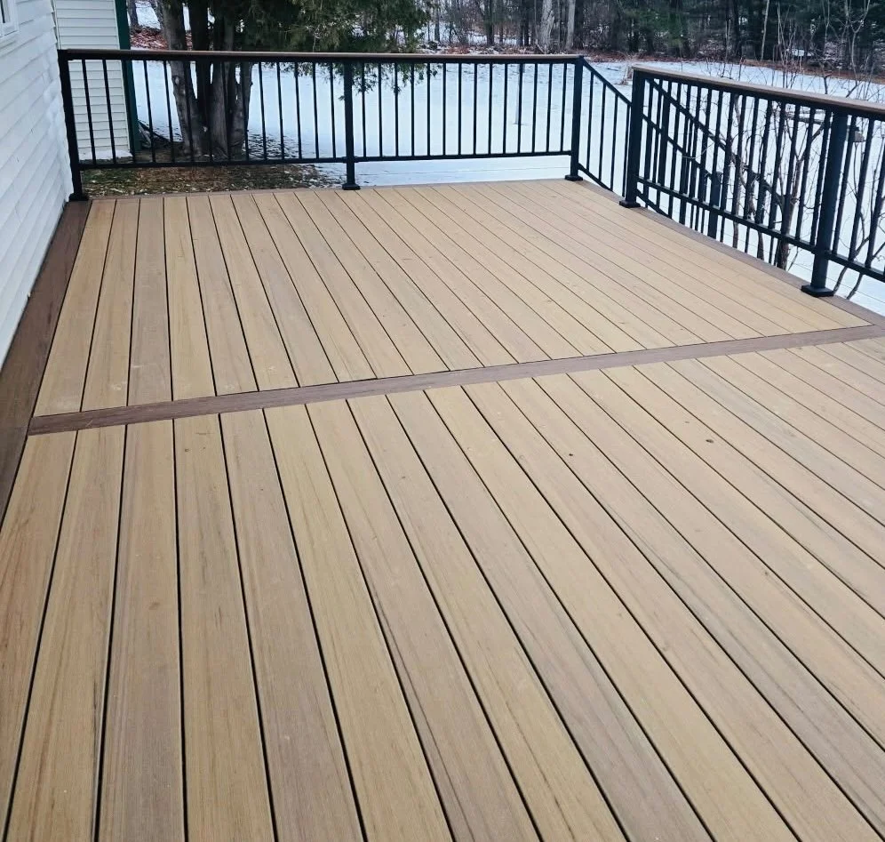 A spacious outdoor wooden deck with a black metal railing, overlooking a snowy landscape with trees and a snow-covered yard.