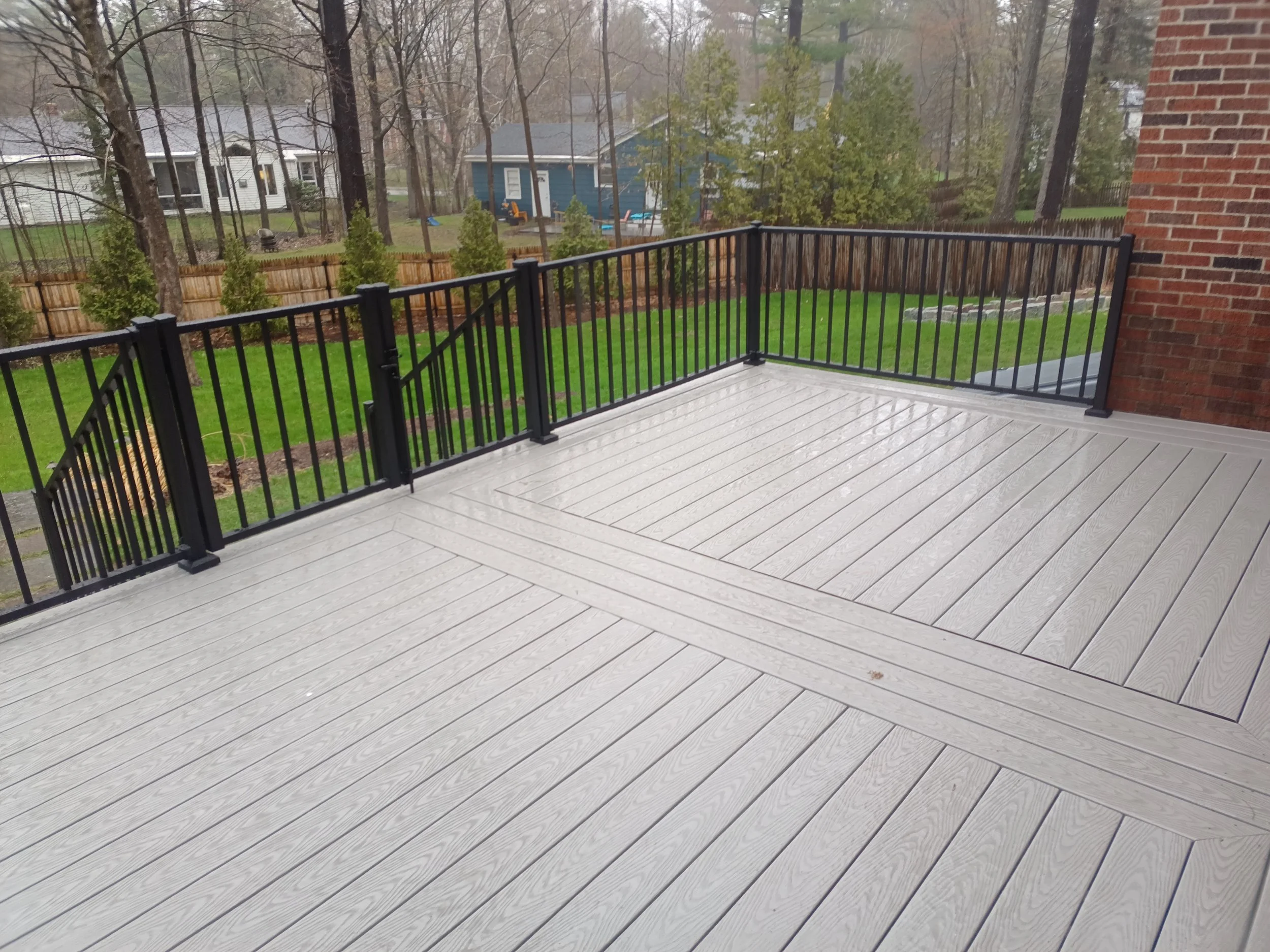 Empty outdoor deck with gray wood floor and black metal railing overlooking a backyard with green grass, trees, and neighboring houses.