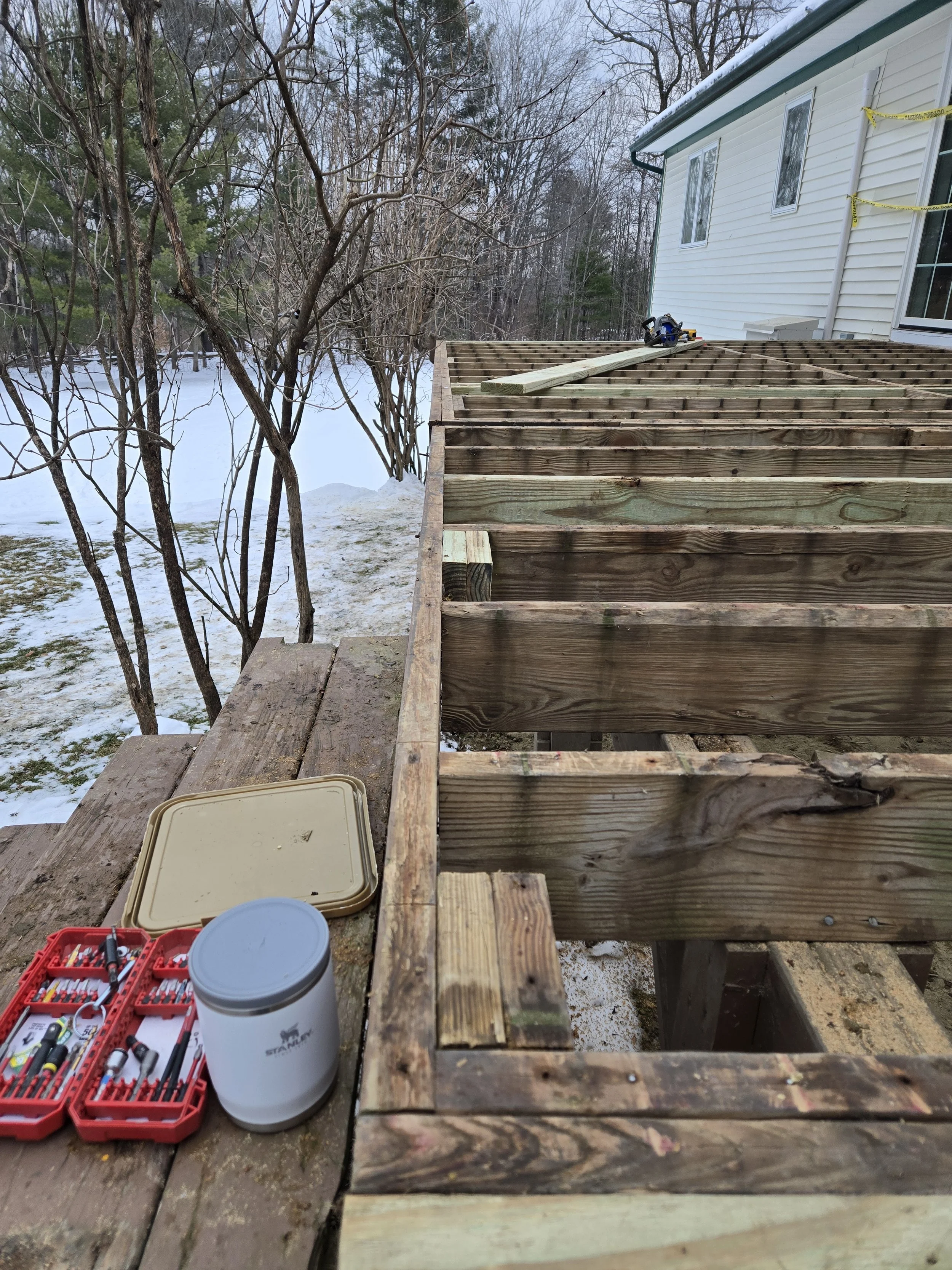 A partially constructed wooden deck attached to a white house, with tools and hardware on a nearby surface, overlooking a snowy yard with leafless trees.