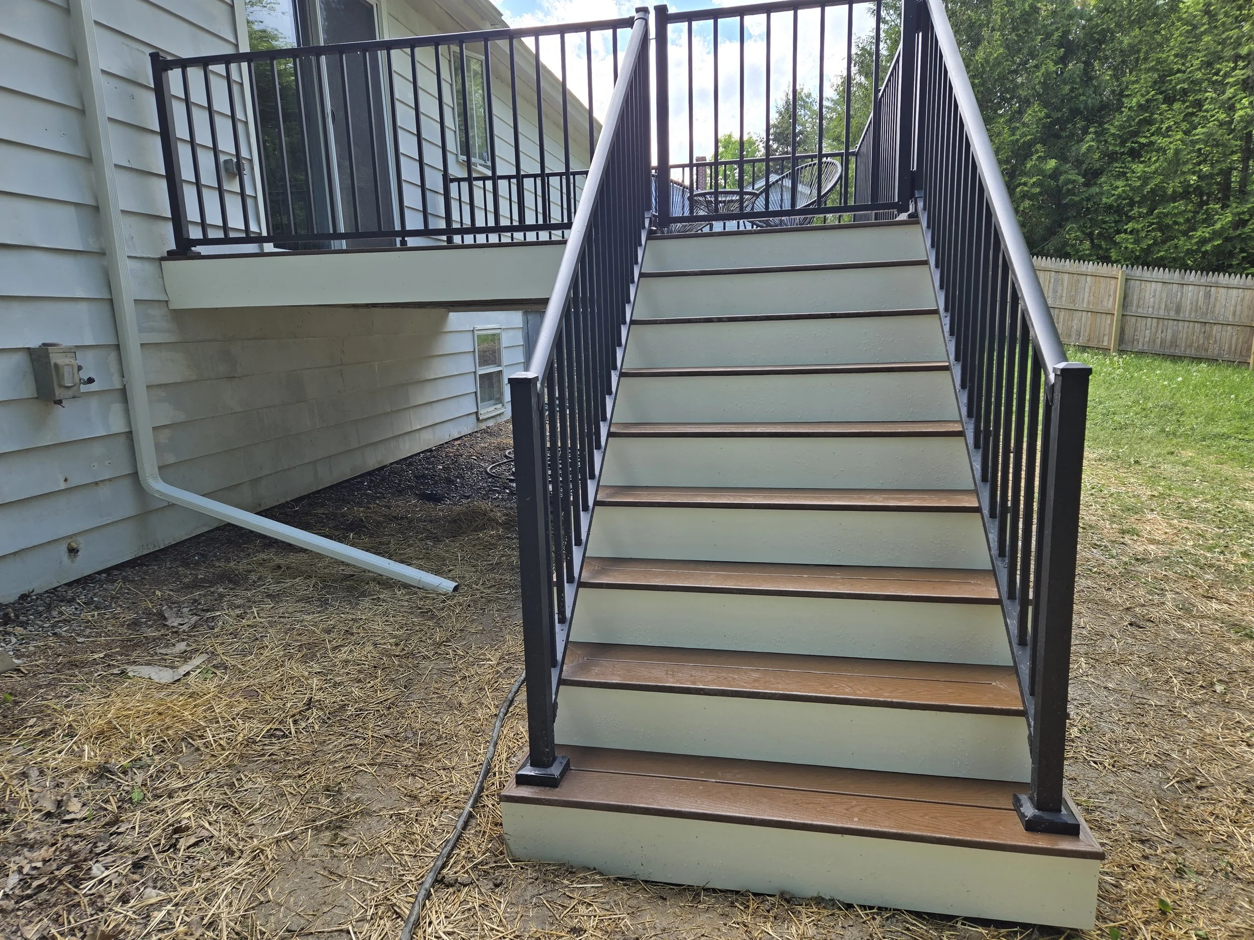 Outdoor staircase with black railings and wooden steps leading to a second-floor deck, attached to a house with beige siding. The yard has dirt and sparse grass, and a wooden fence surrounds the backyard.