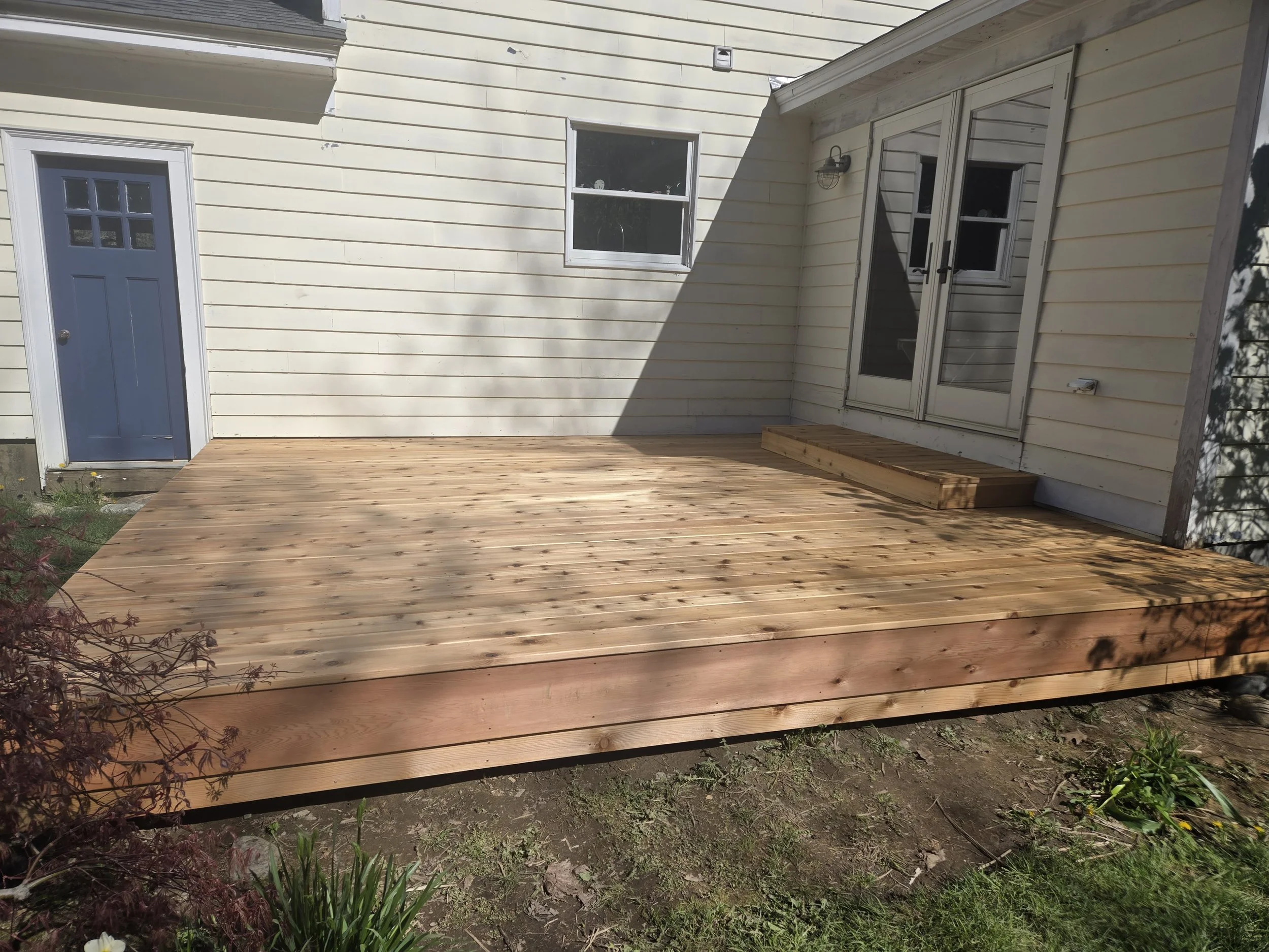 Newly built wooden deck attached to a beige house with a closed glass sliding door and a blue exterior door, surrounded by a garden with plants and soil.