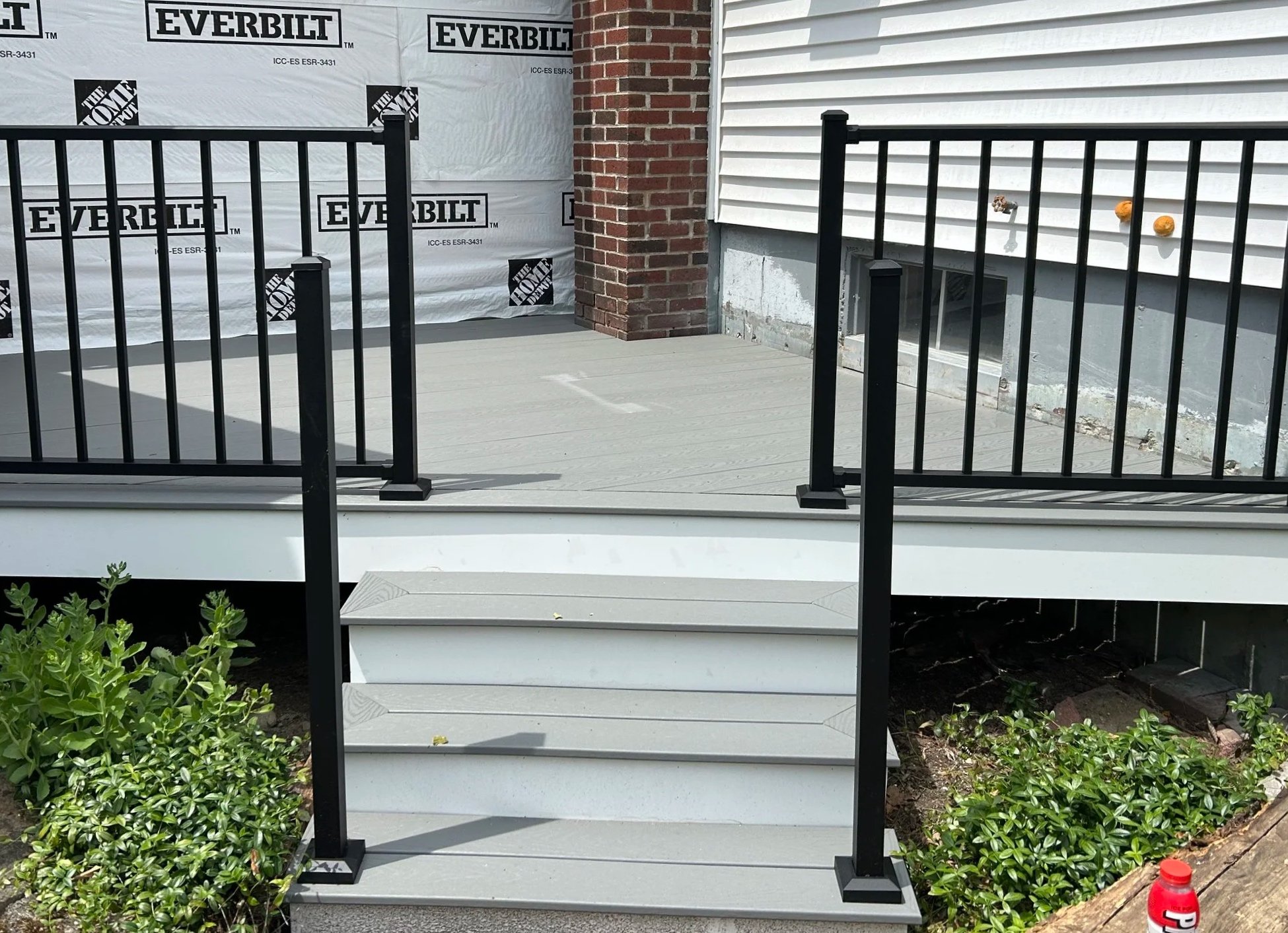 Newly built small porch with black metal railings, light gray steps, and white siding on the house, with some plants and a red bottle in the yard.