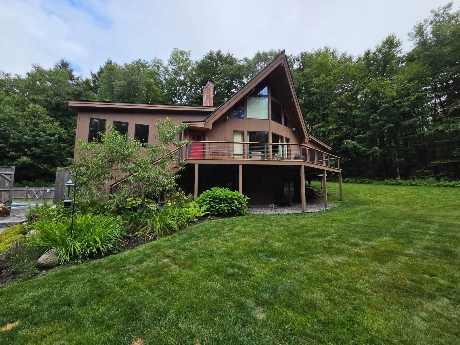 A spacious outdoor deck with a lounge chair, round table with an umbrella, and several chairs, overlooking a green landscape with trees and a cloudy sky.