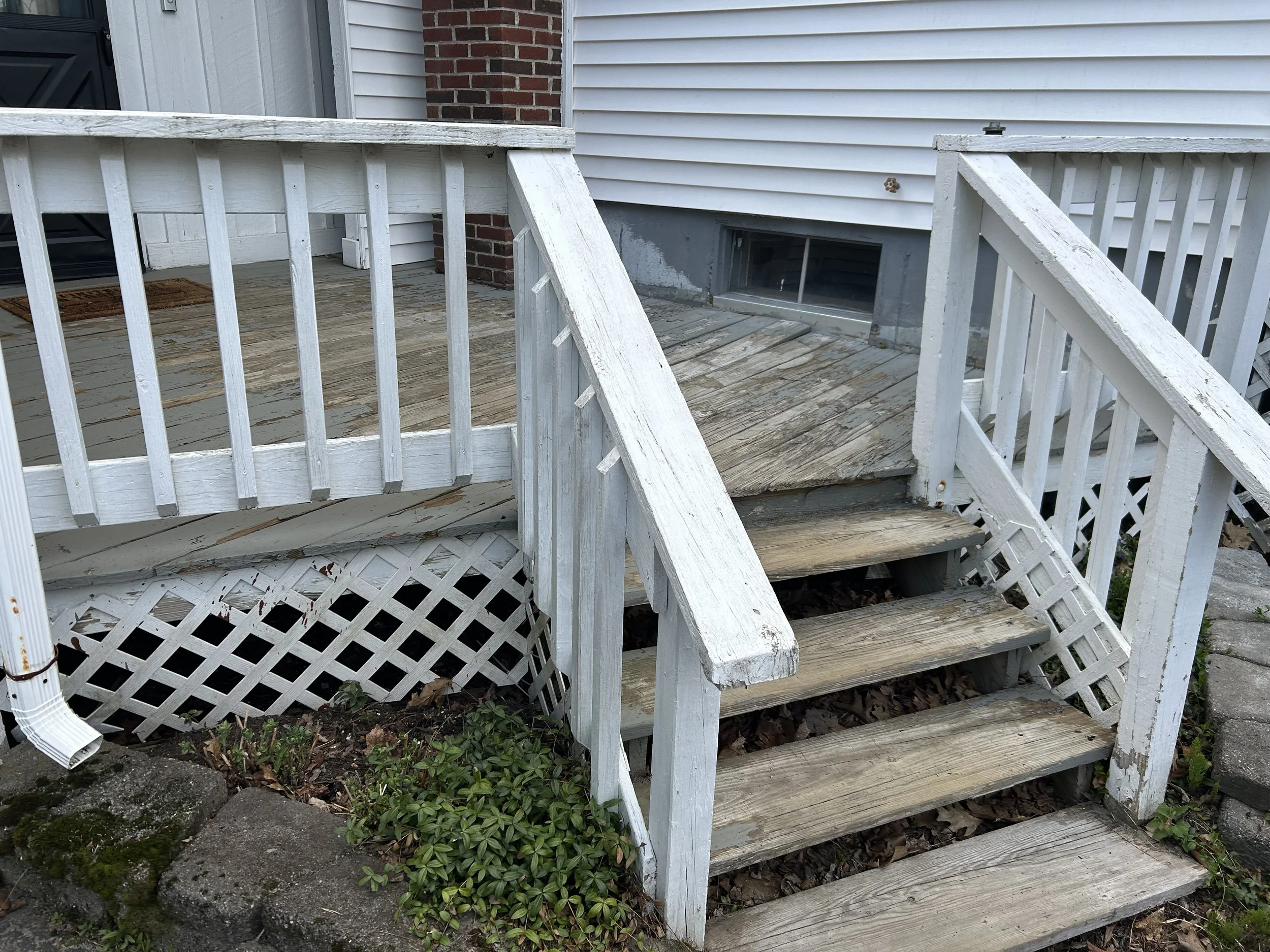 Outdoor wooden porch stairs leading to a deck, with white railings and weathered steps, brown door mat, and a small garden bed with green plants underneath.