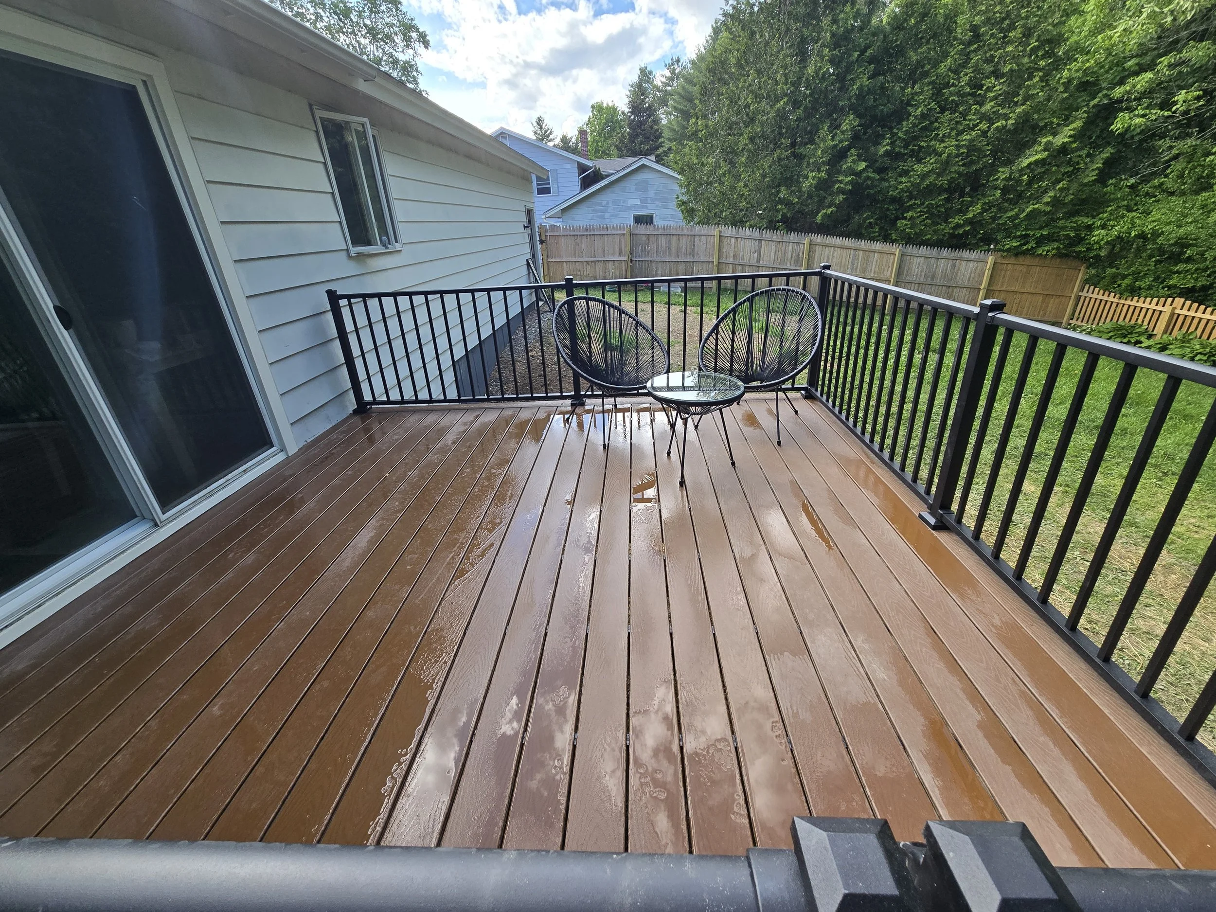 A backyard wooden deck with black metal railing, two black metal chairs, and a glass-top table, with wet surface indicating it recently rained, surrounded by green yard and trees.