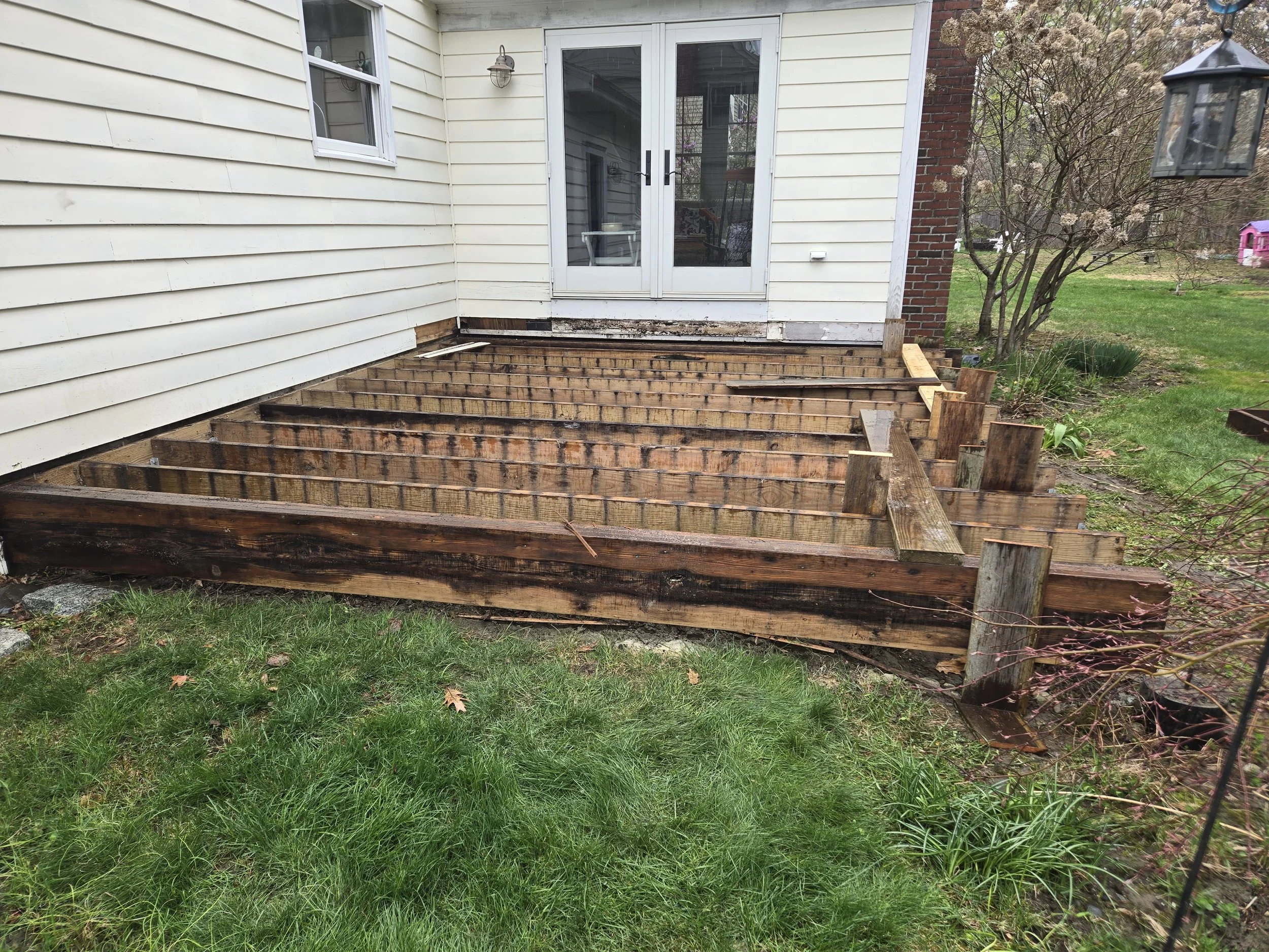Wooden deck framing being constructed outside a house with white siding and a sliding glass door, next to a grassy yard with trees and a lantern hanging from a pole.