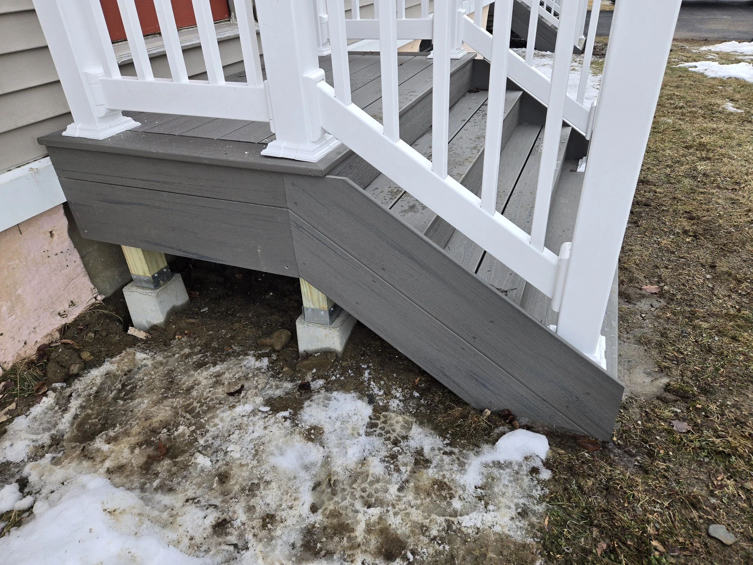 Close-up of a staircase with gray wooden steps and white railing, showing the bottom part of the staircase with supports and visible ground underneath, some snow and dirt on the ground.