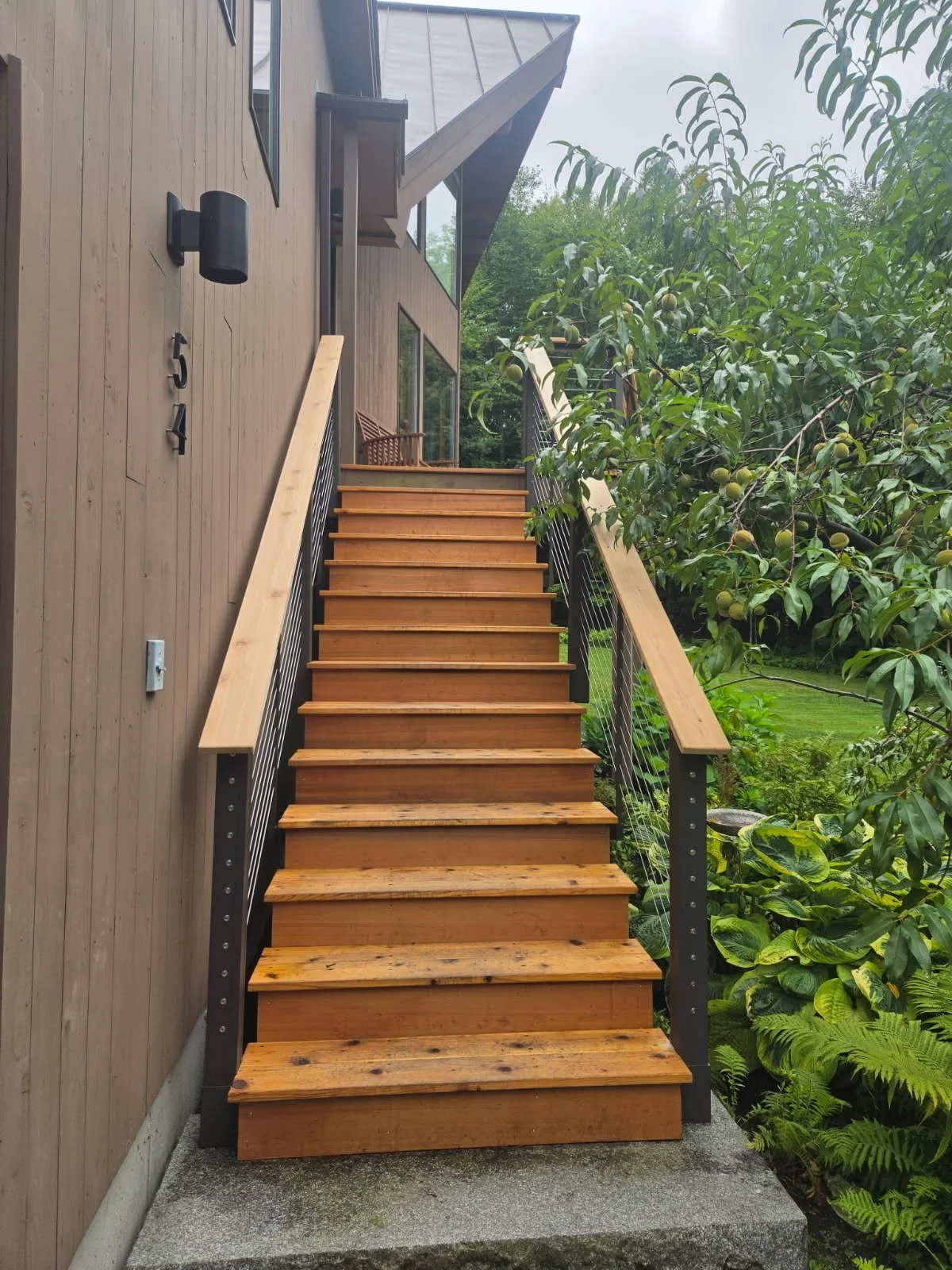 Wooden outdoor staircase leading to a house deck with potted flowers, on a grassy lawn under partly cloudy sky.