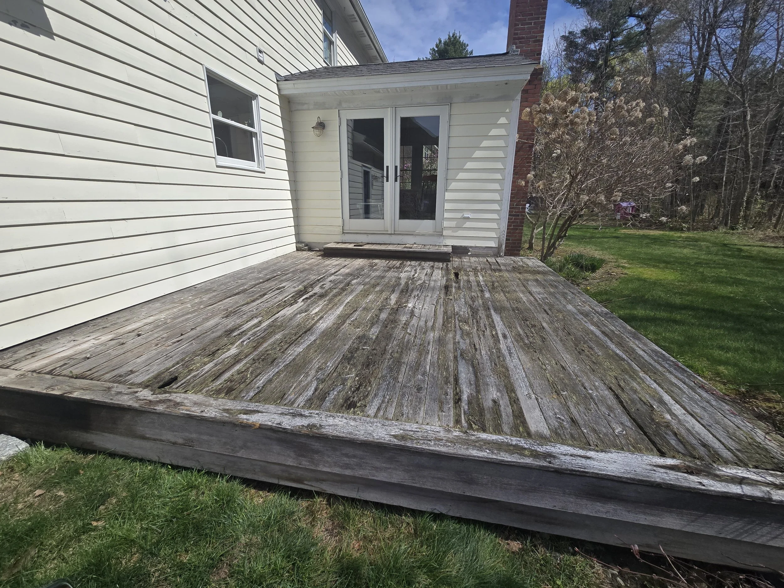 Backyard wooden deck with weathered planks in front of a white house with sliding glass door and window, surrounded by green grass and trees.