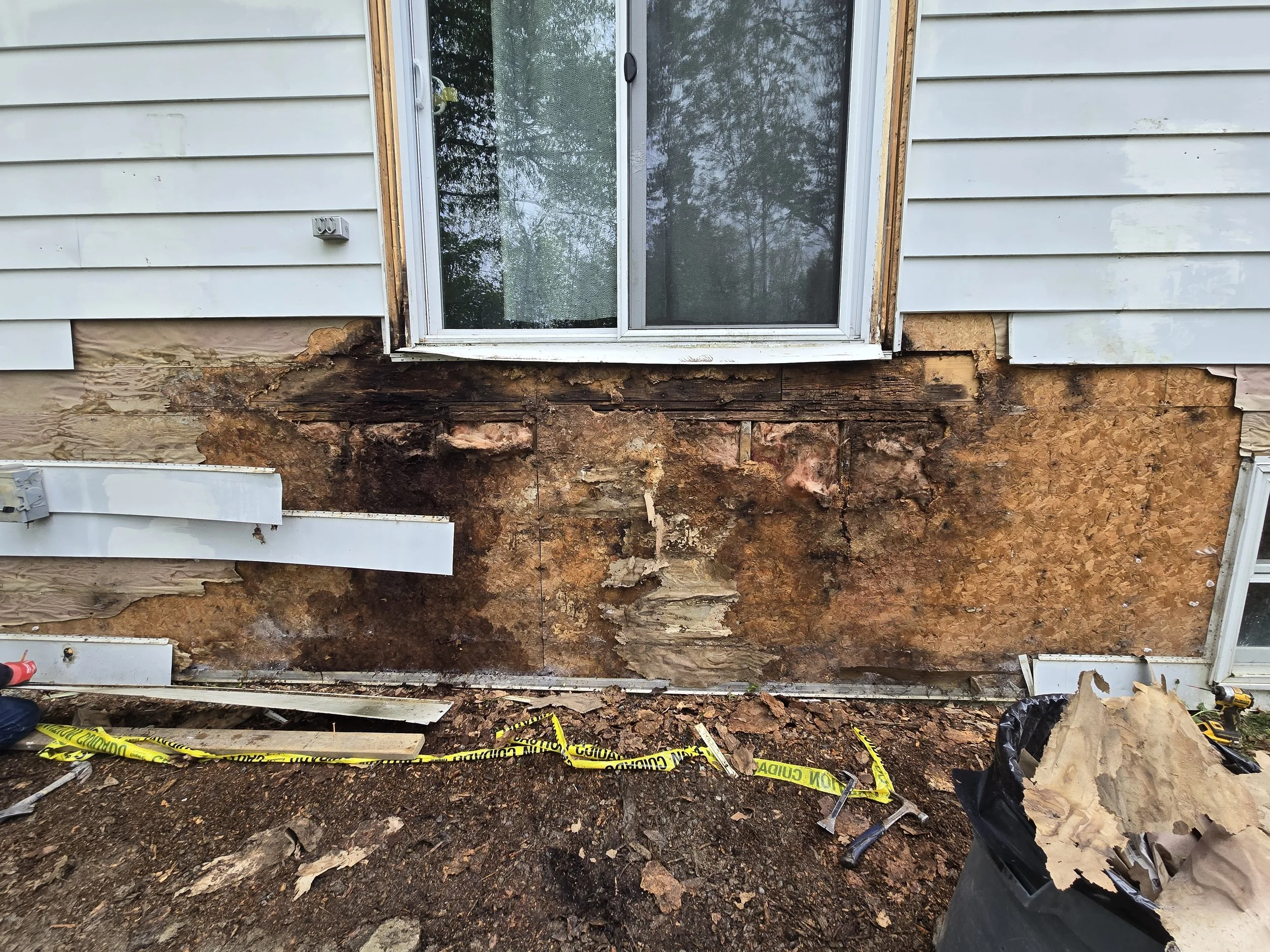 Exterior house wall under repair with damaged siding and exposed wood, construction tools and debris on the ground.