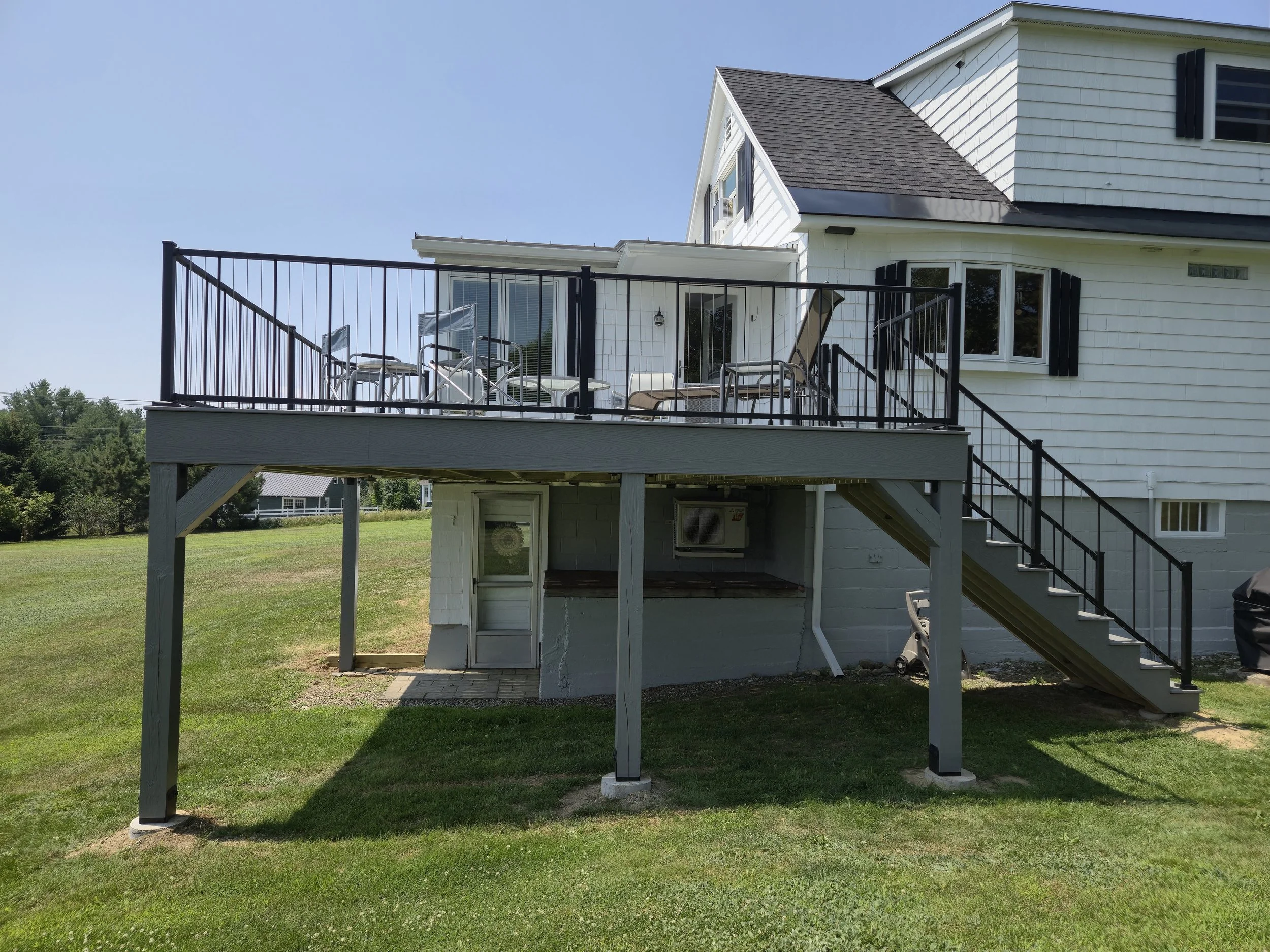 Backyard of a white house with a deck featuring outdoor furniture, stairs leading down to the grass, and surrounded by a green lawn and trees.