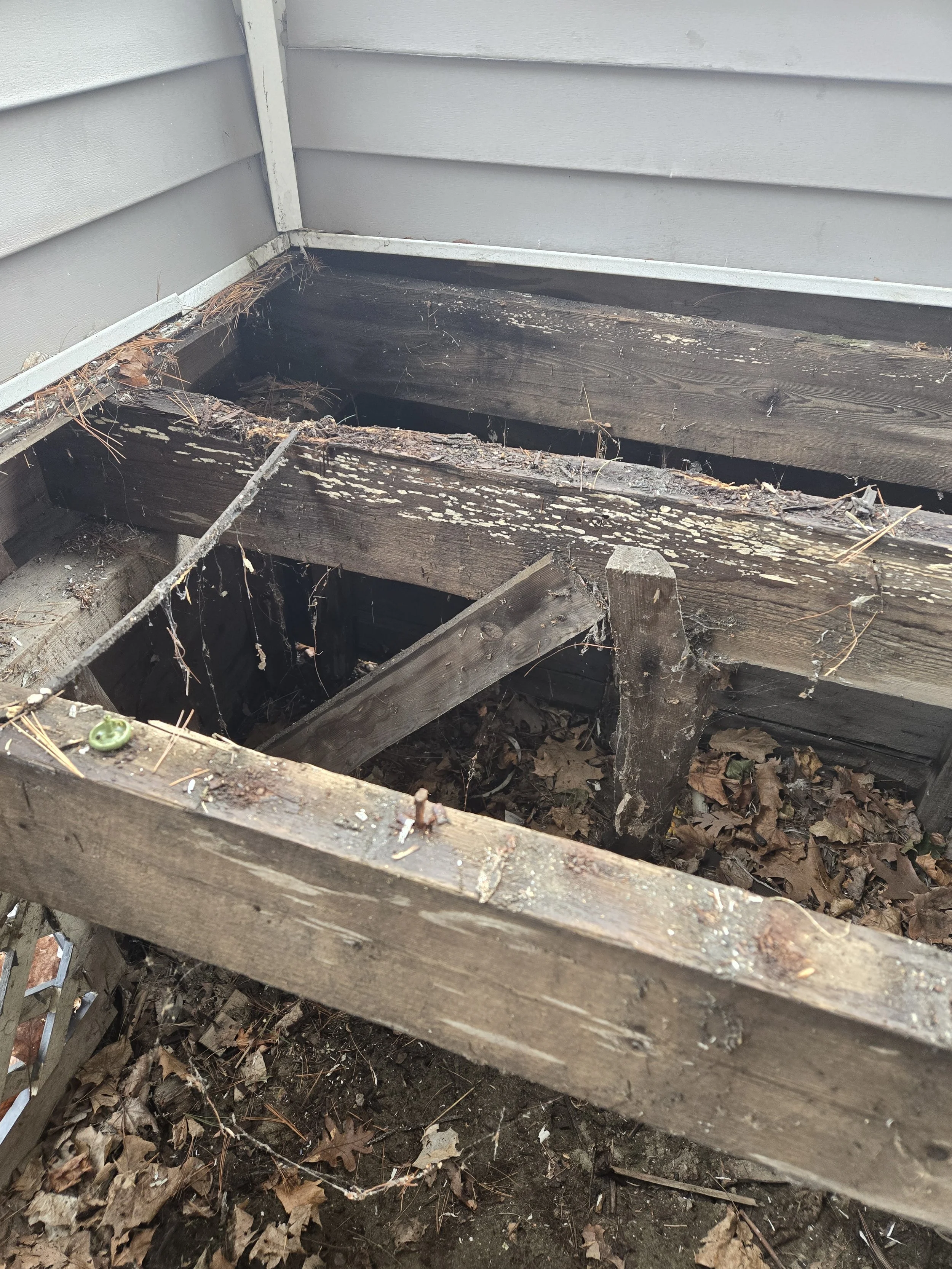 Damaged wooden deck with broken planks, debris, and fallen leaves underneath, next to gray siding of a house.