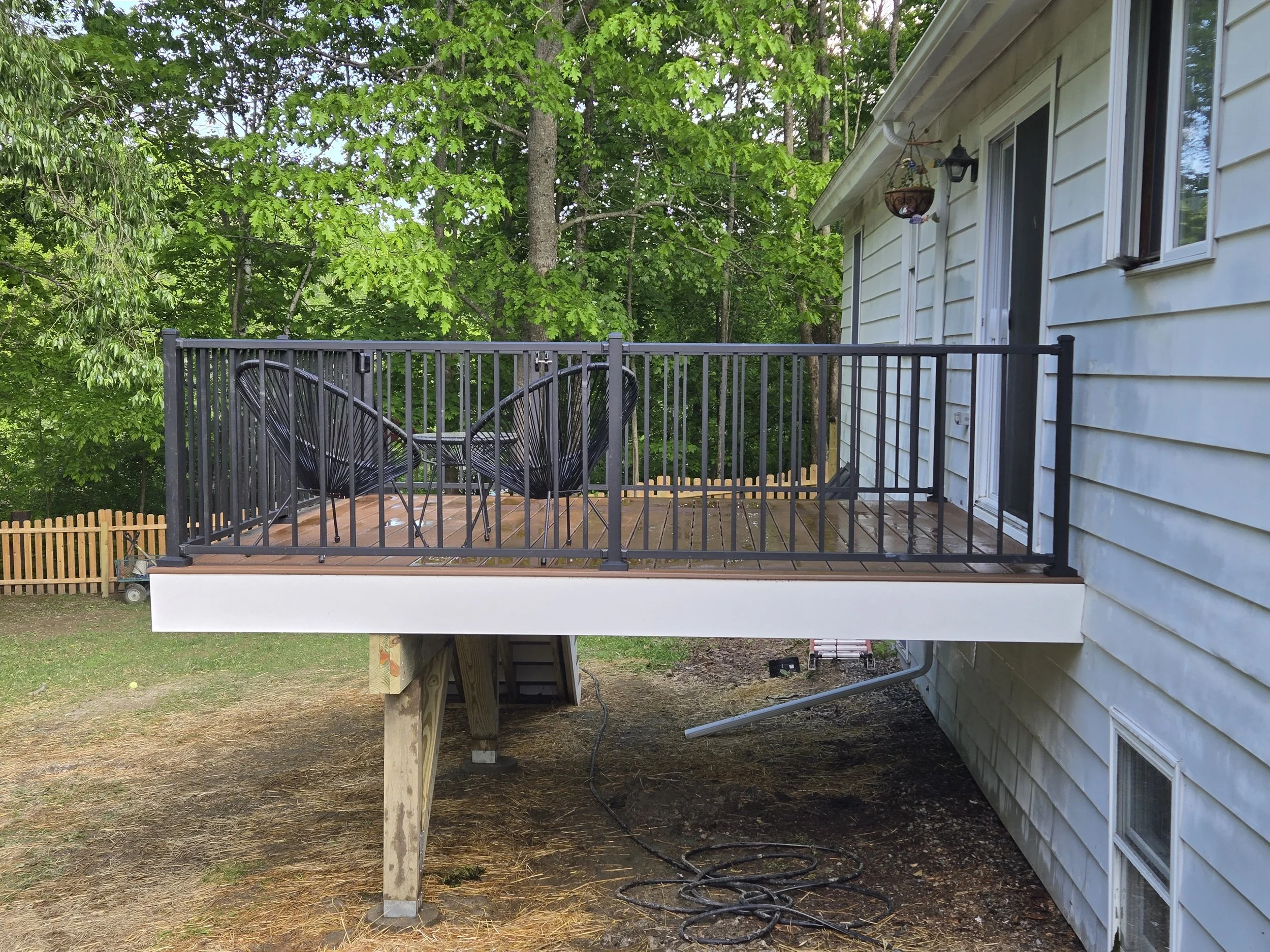 A small outdoor deck with a black metal railing and two black chairs with a small matching table, attached to a white house with horizontal siding, surrounded by green trees.