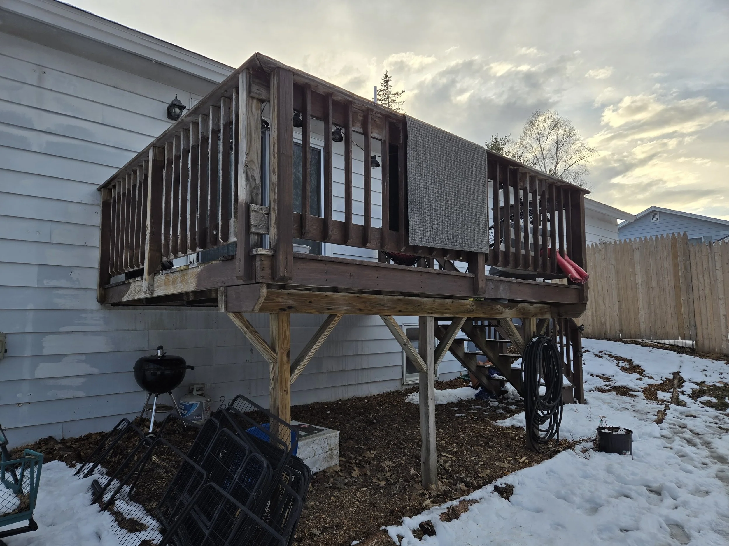 Second-story wooden deck attached to a white house, with stairs leading down, snow on the ground, and a fenced backyard.