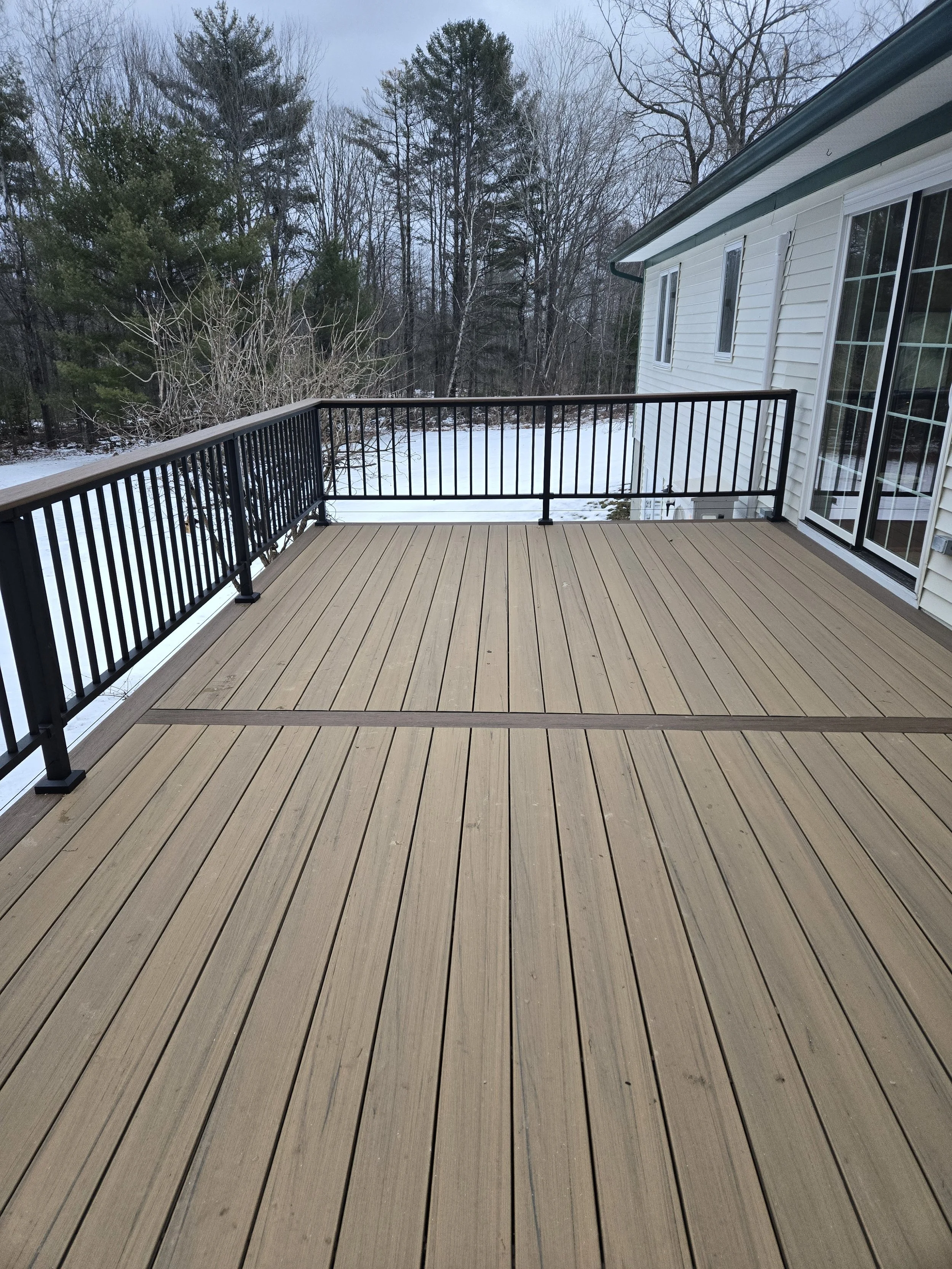 A wooden deck attached to a house with a black metal railing, overlooking a snowy backyard with leafless trees in the background.