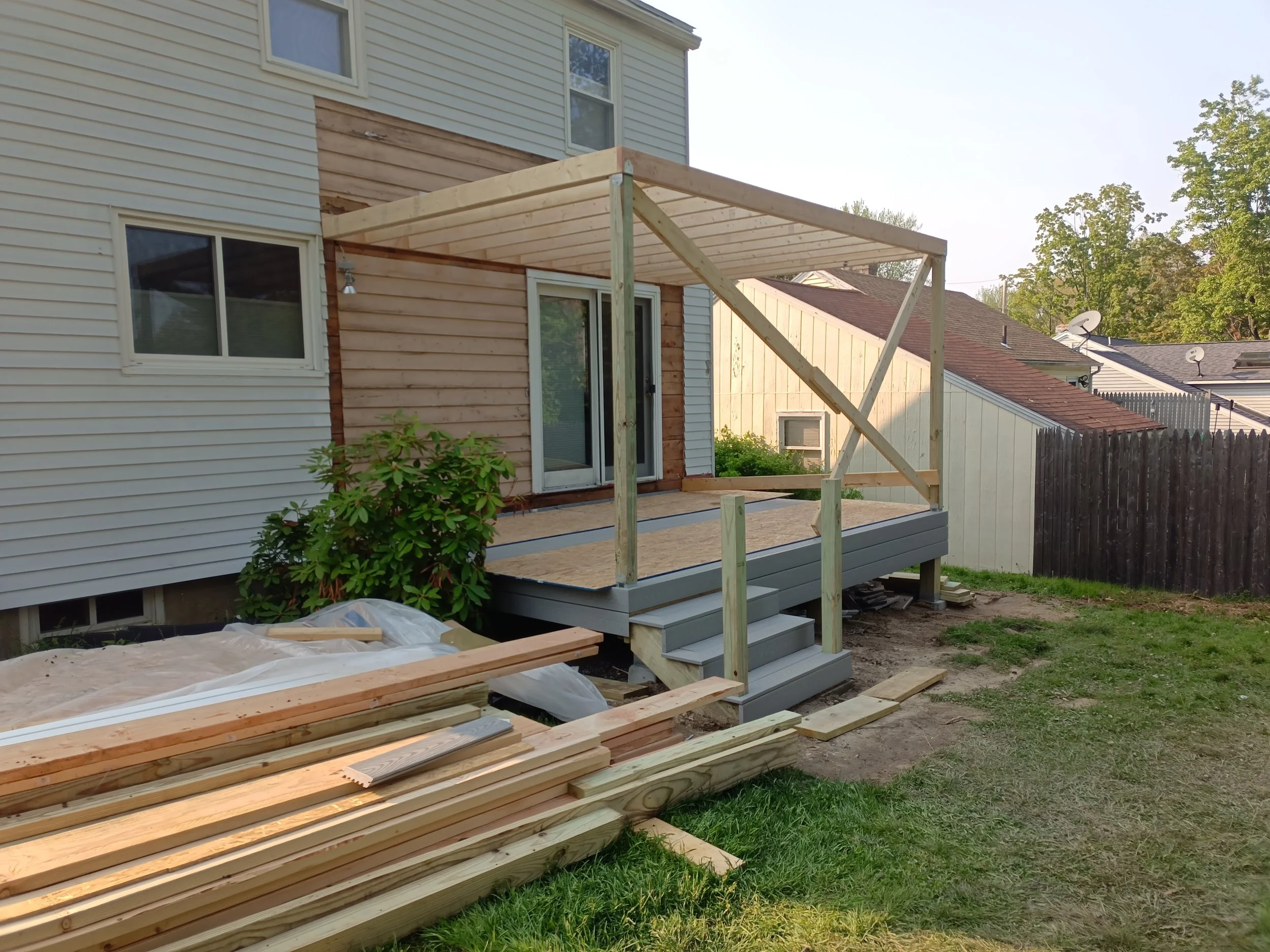 Backyard of a house under construction with a new wooden deck and porch roof in progress. Building materials and stairs are visible. Fencing and neighboring houses in the background.