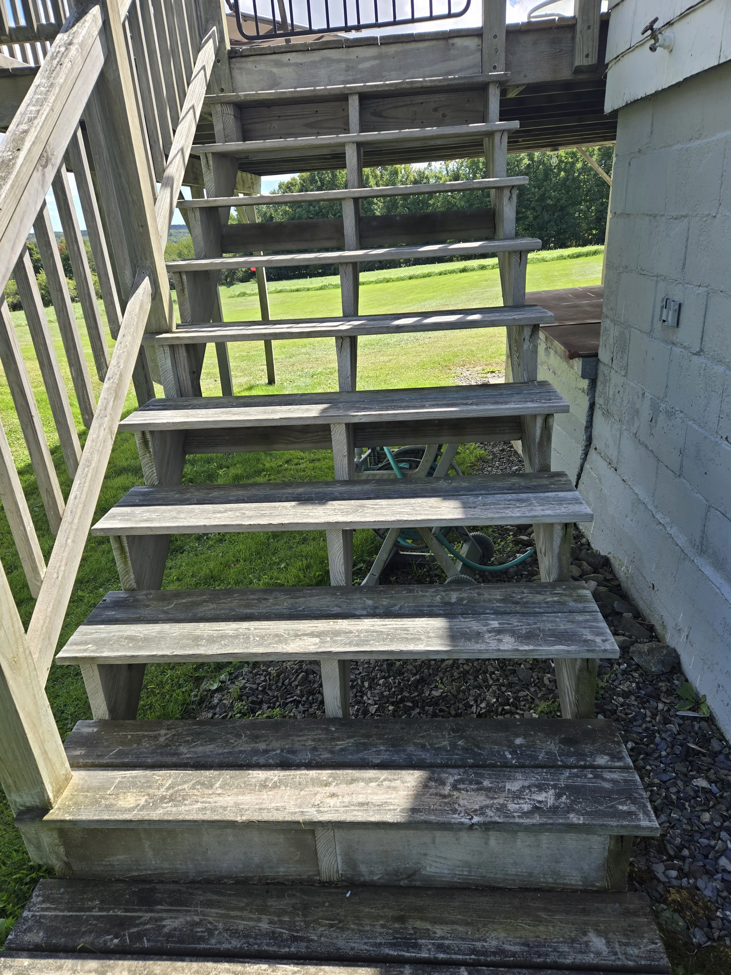 Wooden outdoor staircase leading from a deck to the ground, with grass and trees in the background.