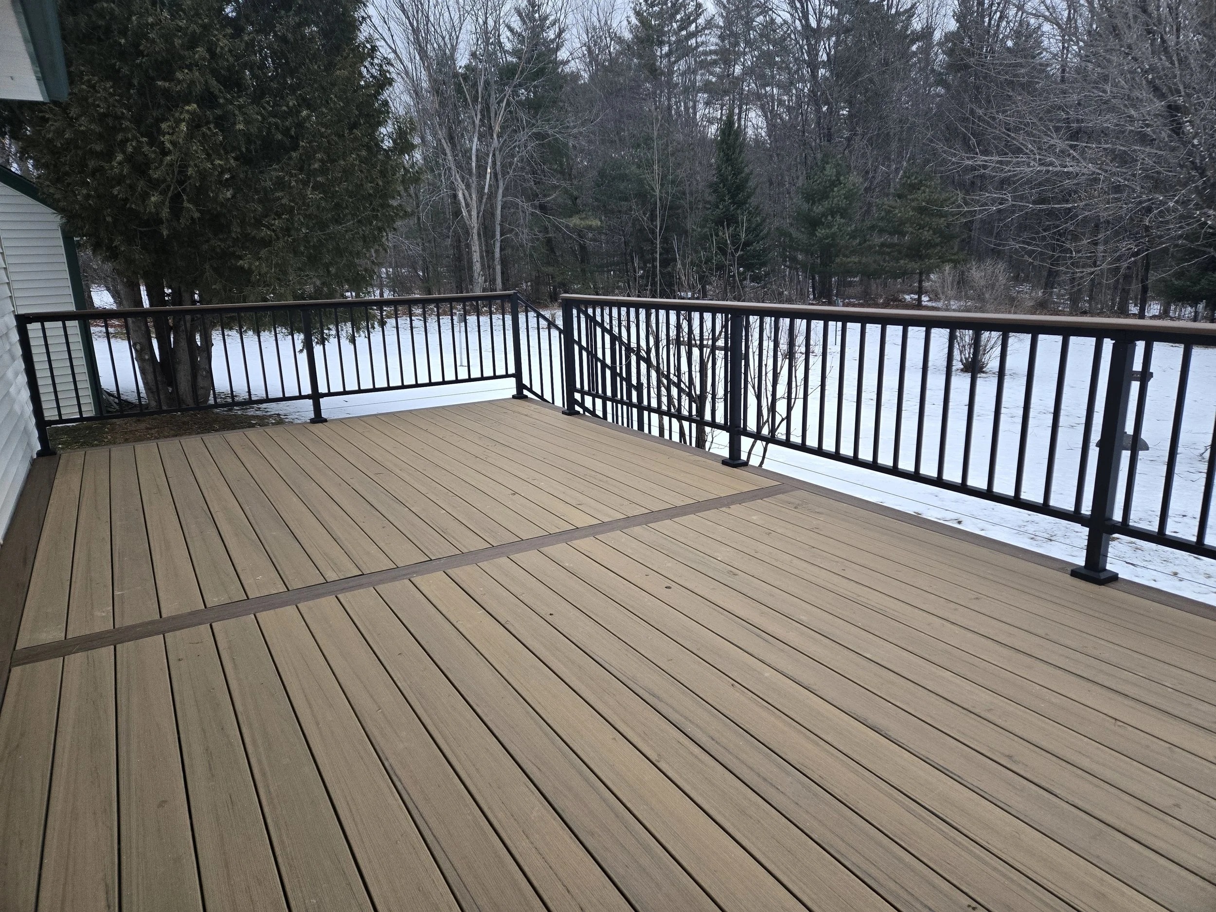 Empty wooden deck with black metal railing overlooking a snow-covered backyard with trees.