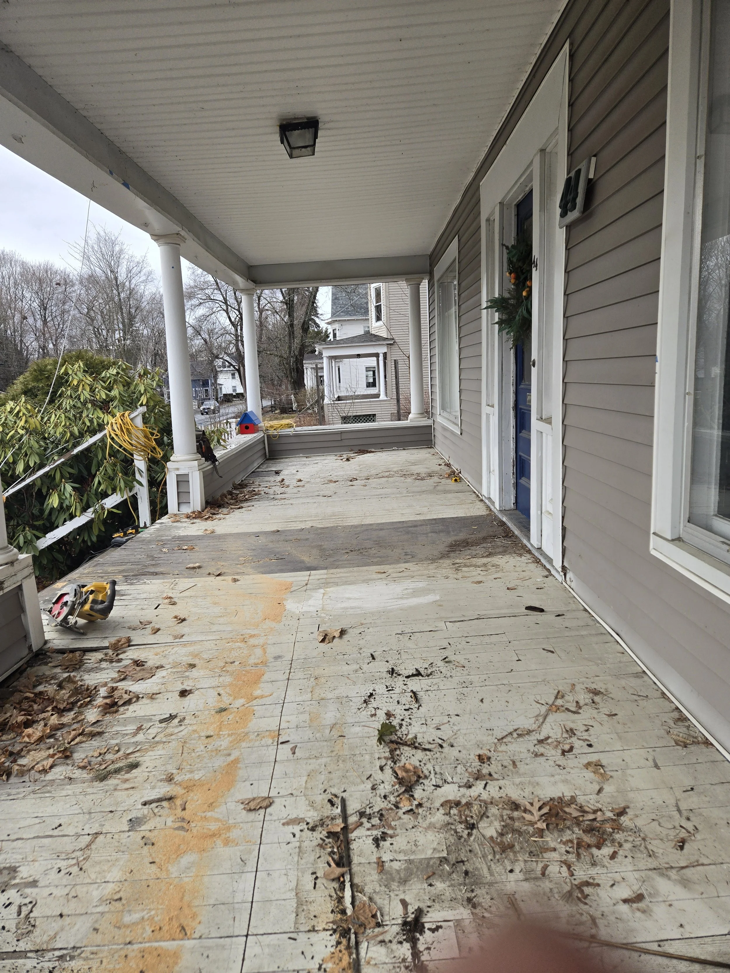 Empty front porch with peeling paint, scattered leaves, yard tools, and a wreath on the door, with neighboring houses and bare trees in the background.