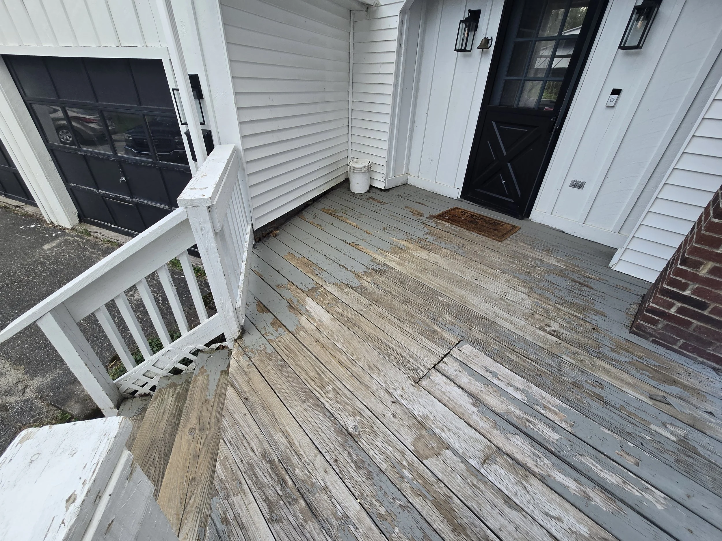 Front porch with weathered wooden floor, black door, white siding, and a small white bucket near the corner.