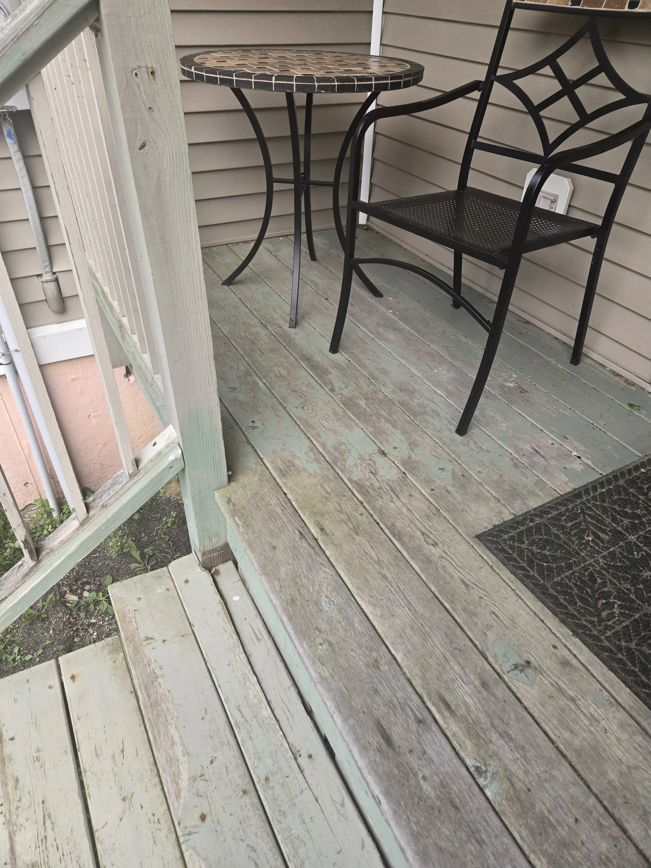 A small outdoor wooden porch with a black metal chair and a small round table with a mosaic top, next to beige siding and a doormat.