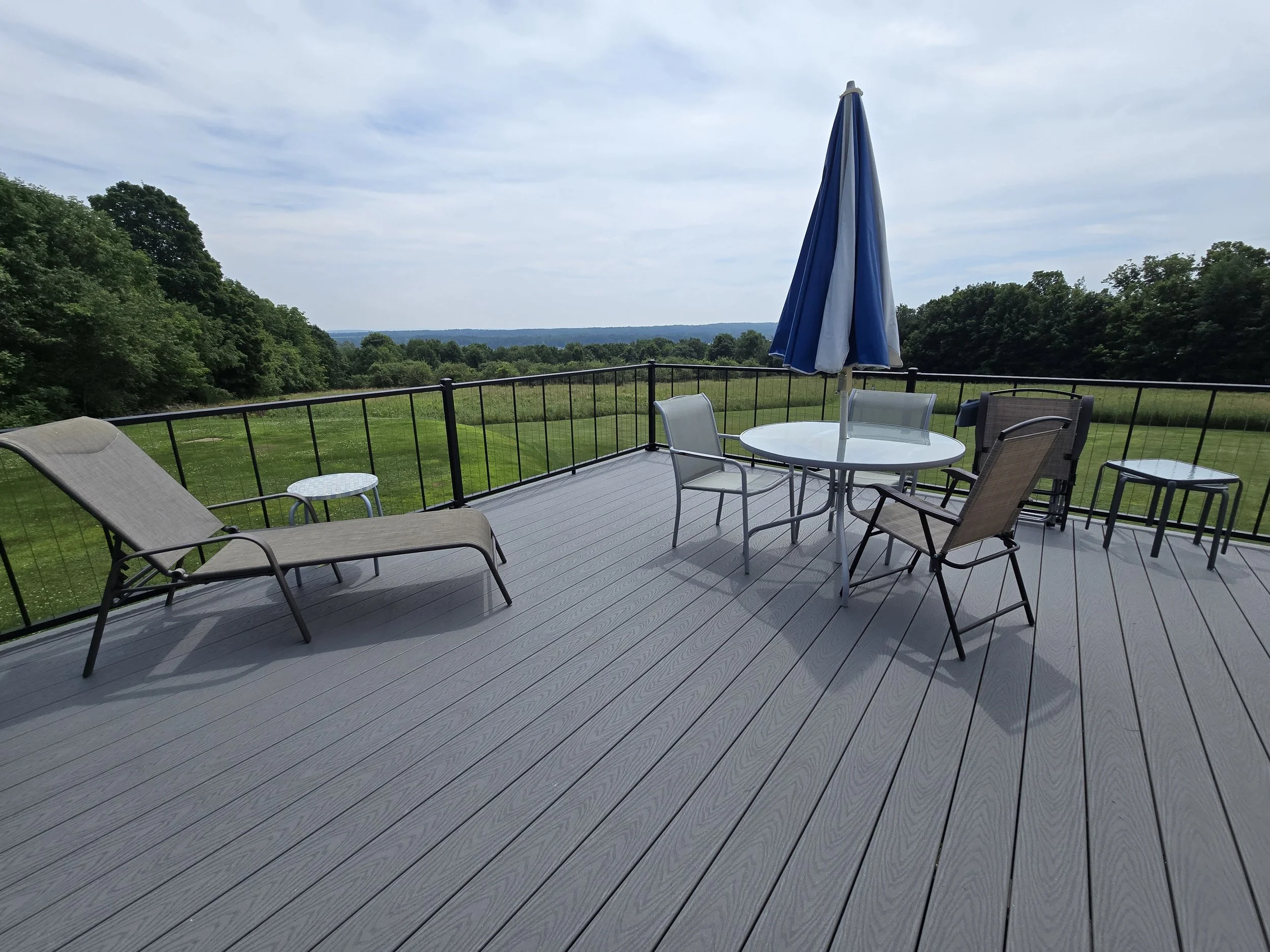 A spacious outdoor deck with a lounge chair, round table with an umbrella, and several chairs, overlooking a green landscape with trees and a cloudy sky.