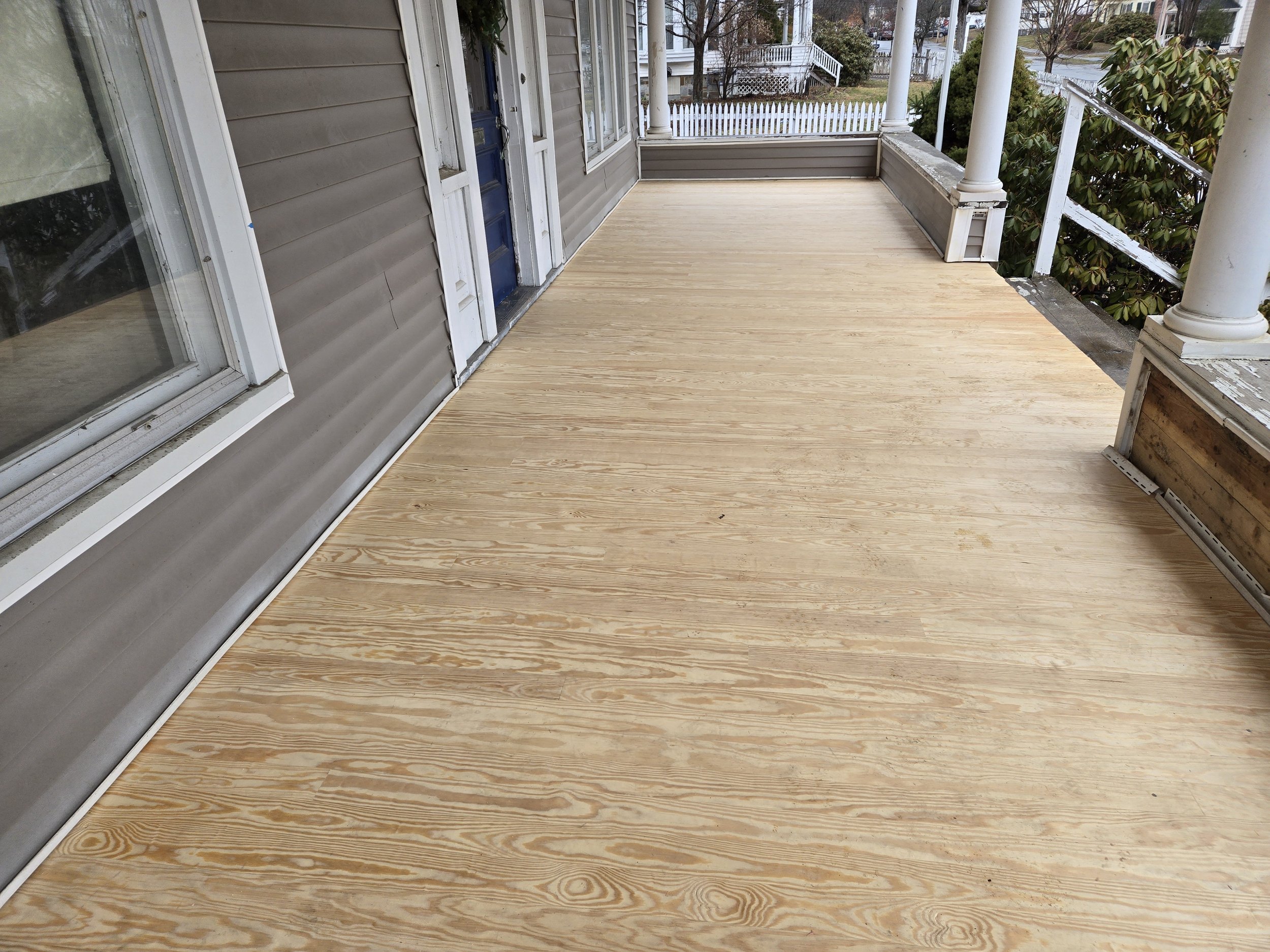 A wooden porch deck outside a house with gray siding, white trim, and white columns. There is a blue door and a glass sliding door. In the background, there are trees, shrubs, and neighboring houses.