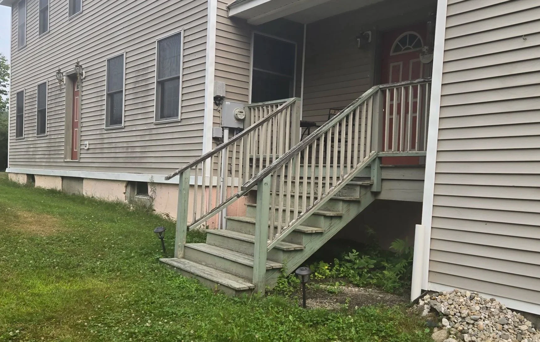 Exterior view of a beige multi-unit apartment building with stairs leading to a porch. The building has multiple windows, some with curtains, and two red doors. There are small landscaped areas with grass, rocks, and outdoor lighting.