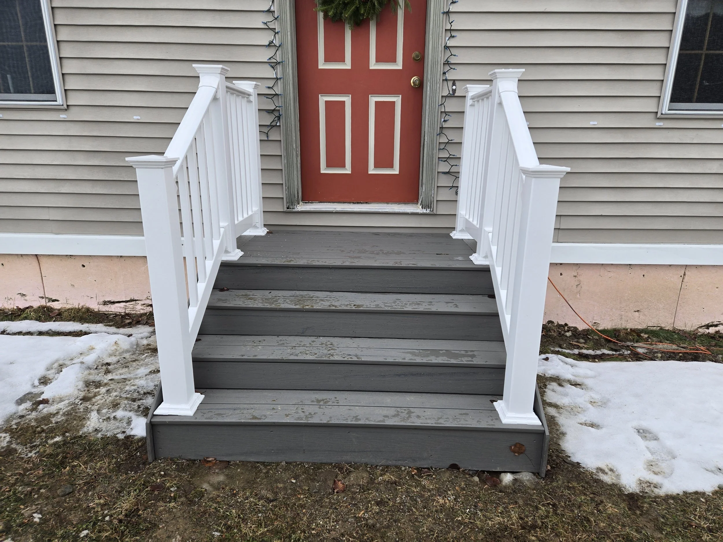 Front porch with gray wooden stairs, white railing, and a red door with windows, surrounded by beige siding and snow on the ground.