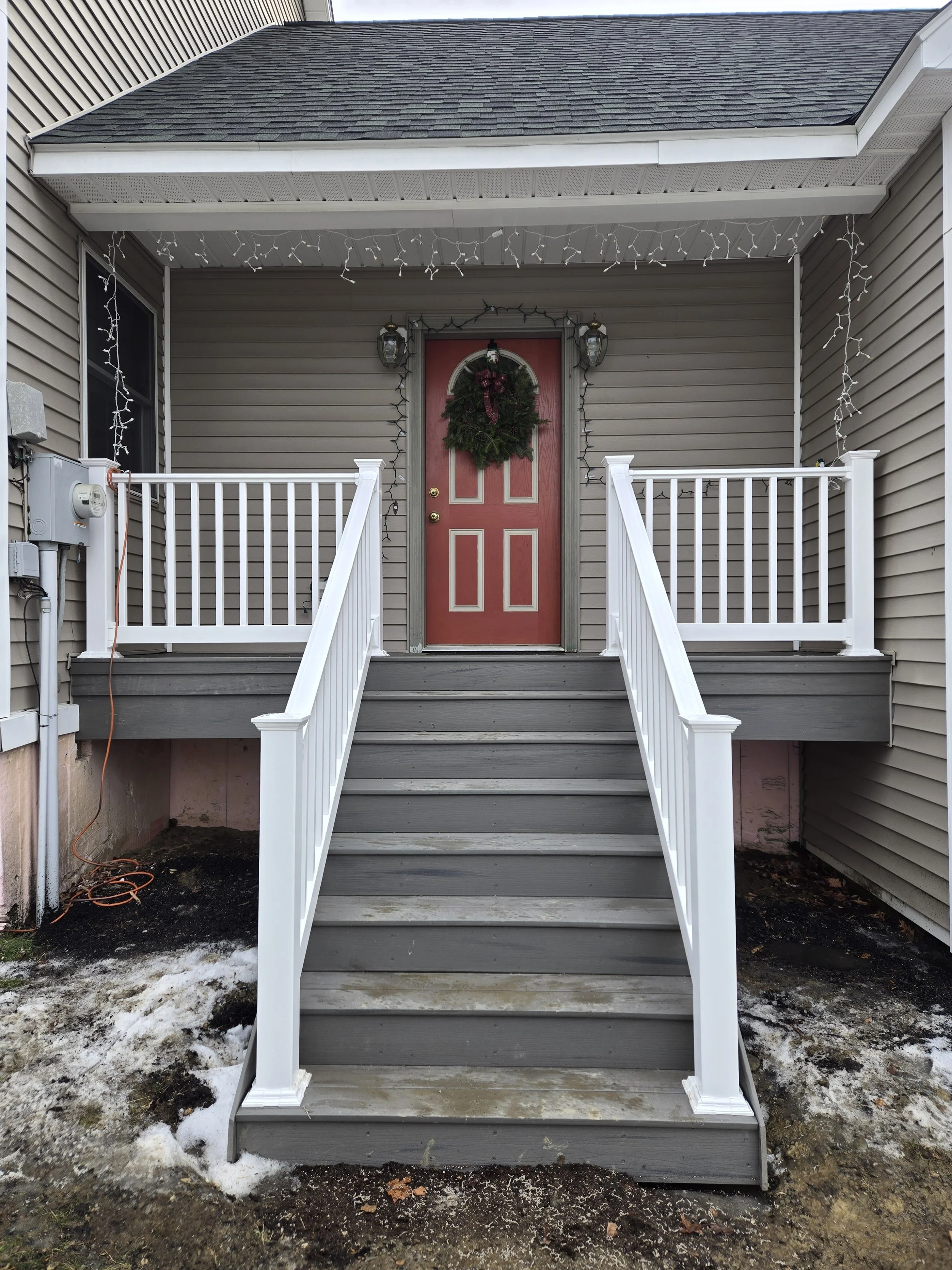 Front porch of a house with a red door decorated with a Christmas wreath, string lights, and a small set of stairs leading to the entrance.