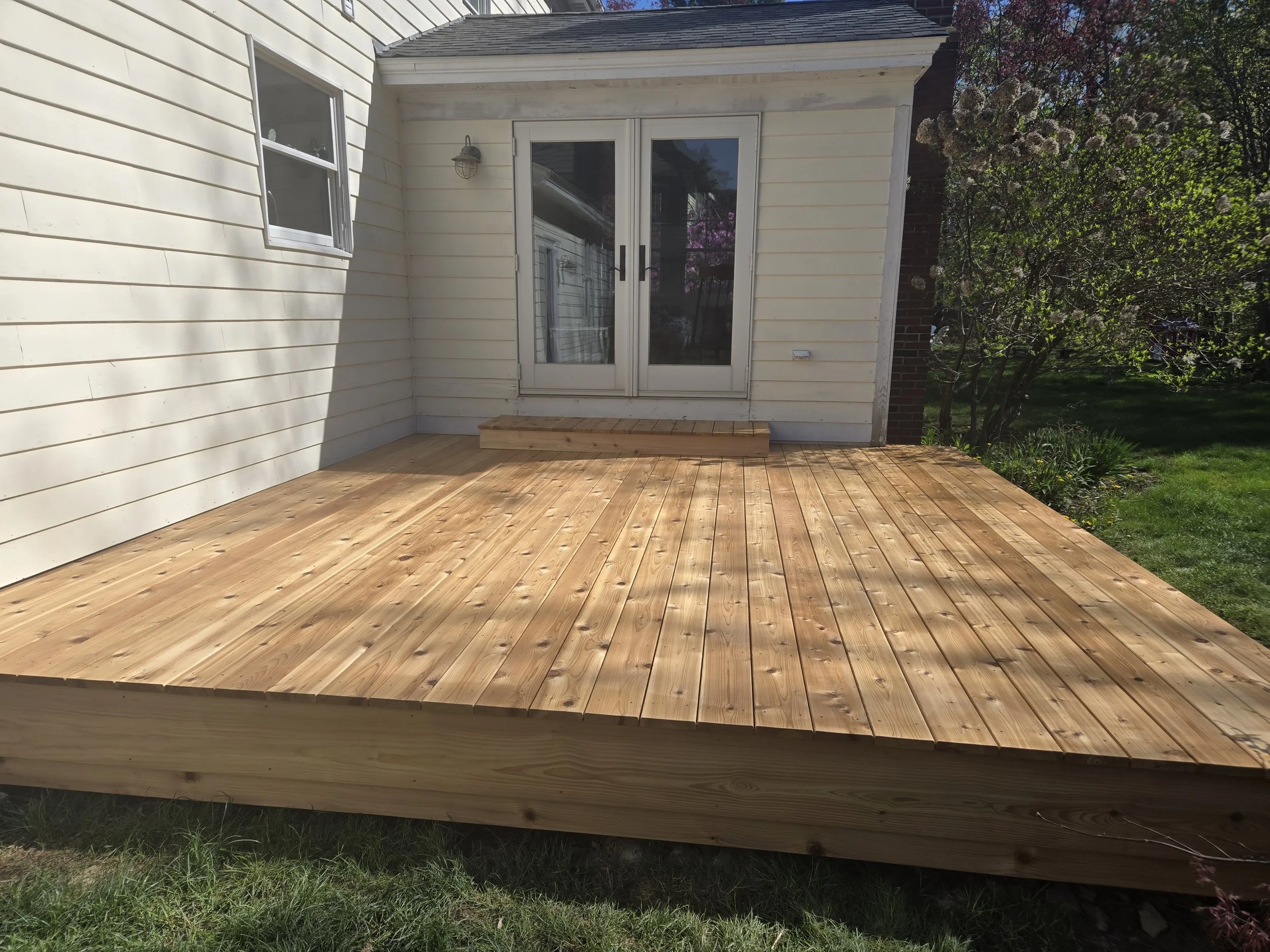 Newly constructed wooden deck attached to the back of a house, with double sliding glass doors leading into the interior. The house has white siding, and there is a small step at the entrance to the deck. The yard beyond the deck is lush with green grass and flowering bushes.