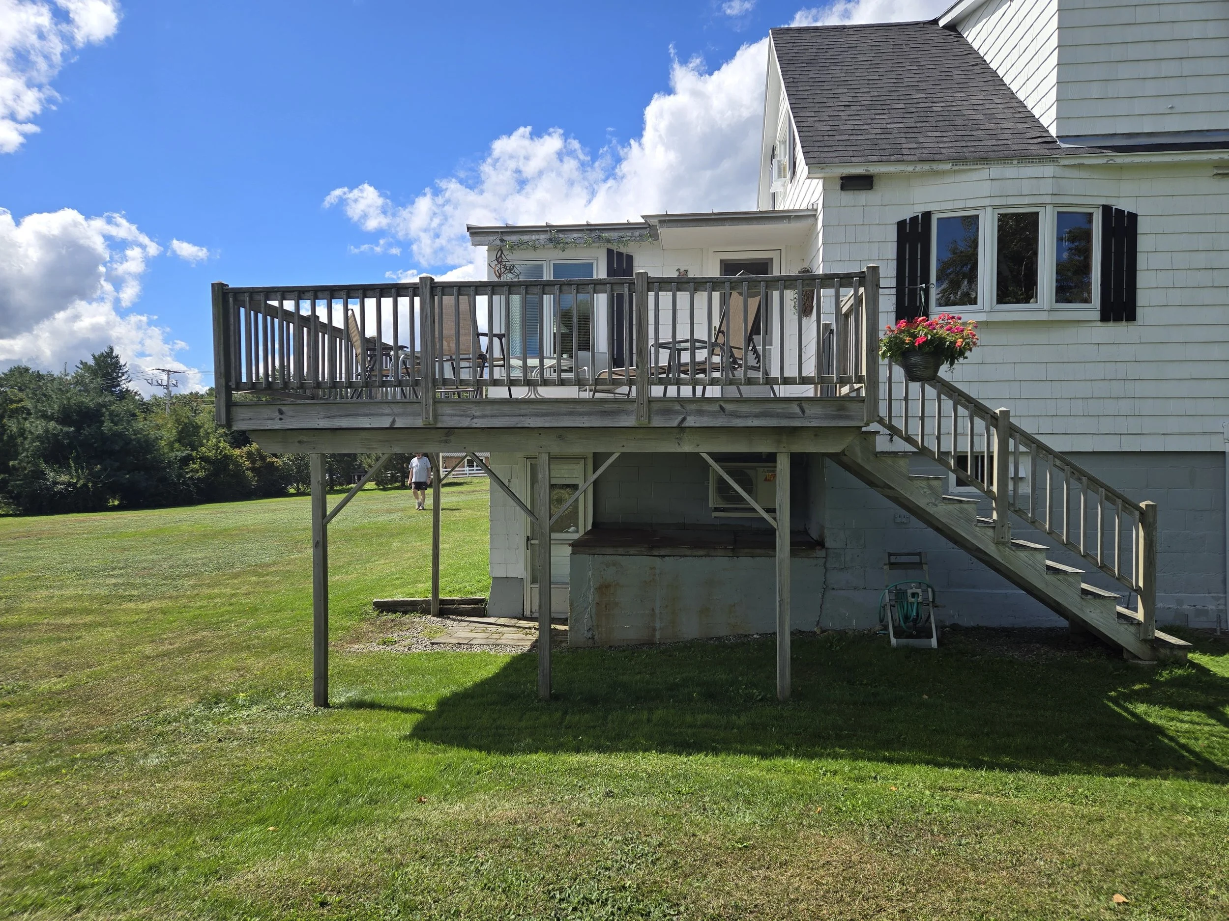 Backyard with a wooden raised deck attached to a white house, featuring a staircase, outdoor furniture, and hanging flower basket, on a sunny day with a blue sky and green lawn.