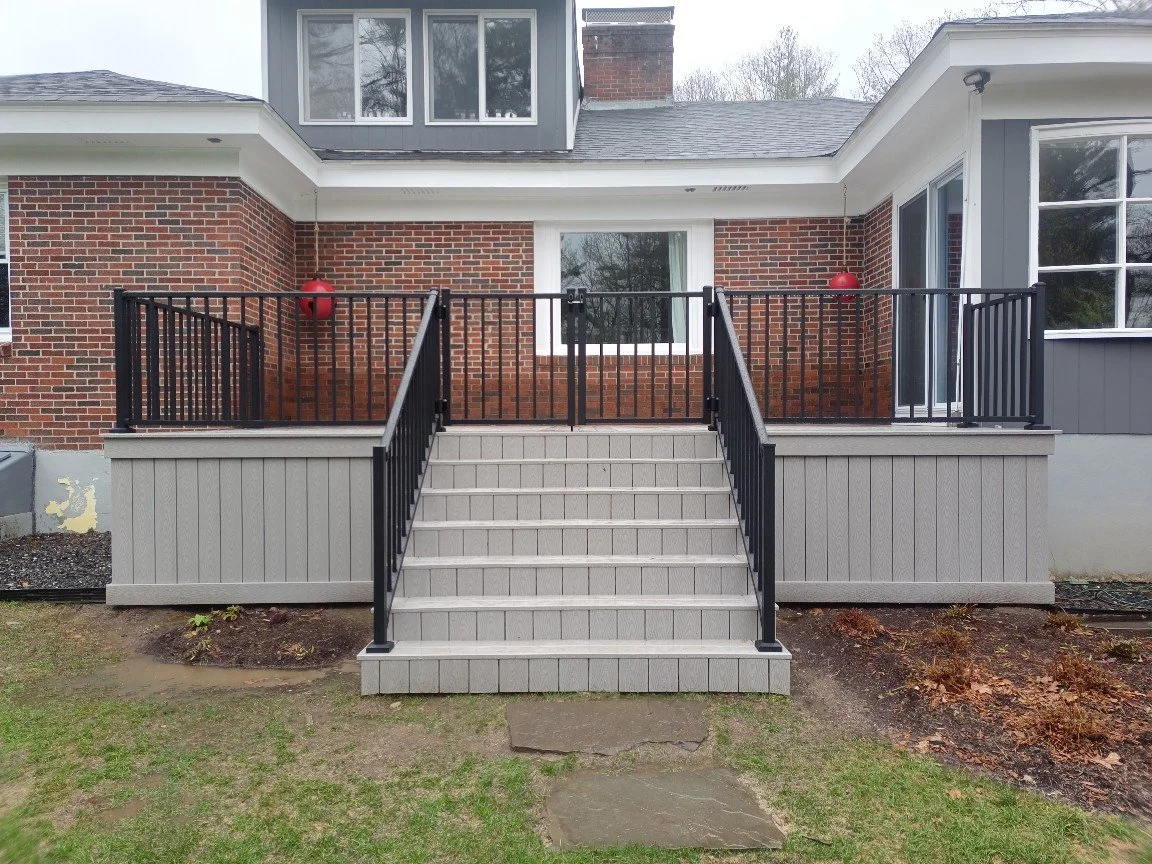 Backyard deck with stairs, black railing, brick house, and large windows.