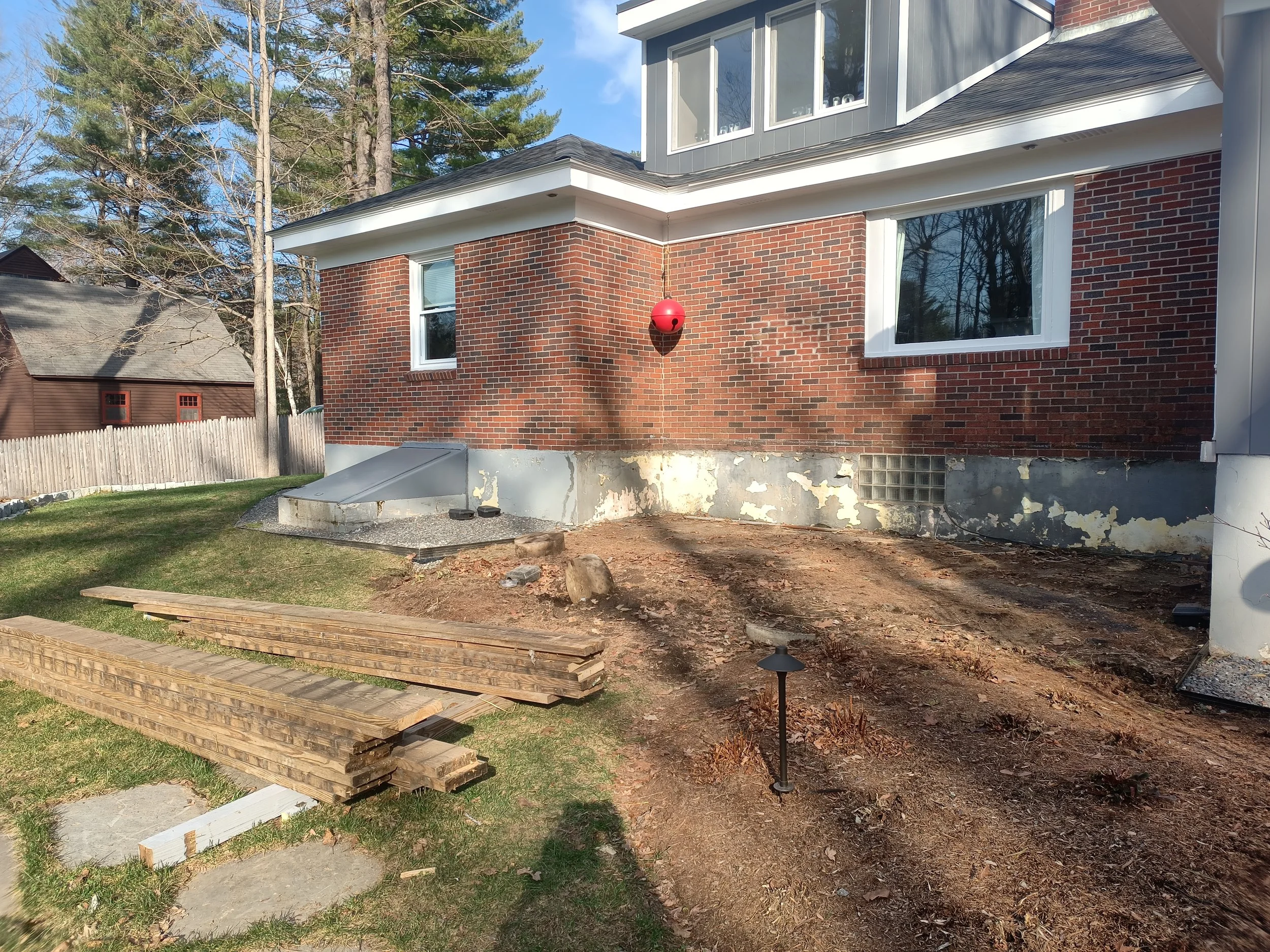 Backyard of a house under construction or renovation, with exposed dirt and wood planks on the ground, a small grassy area, and a brick house with white-framed windows. There is a red spherical object on the house wall, small outdoor lights along the ground, and a fenced yard with trees in the background.