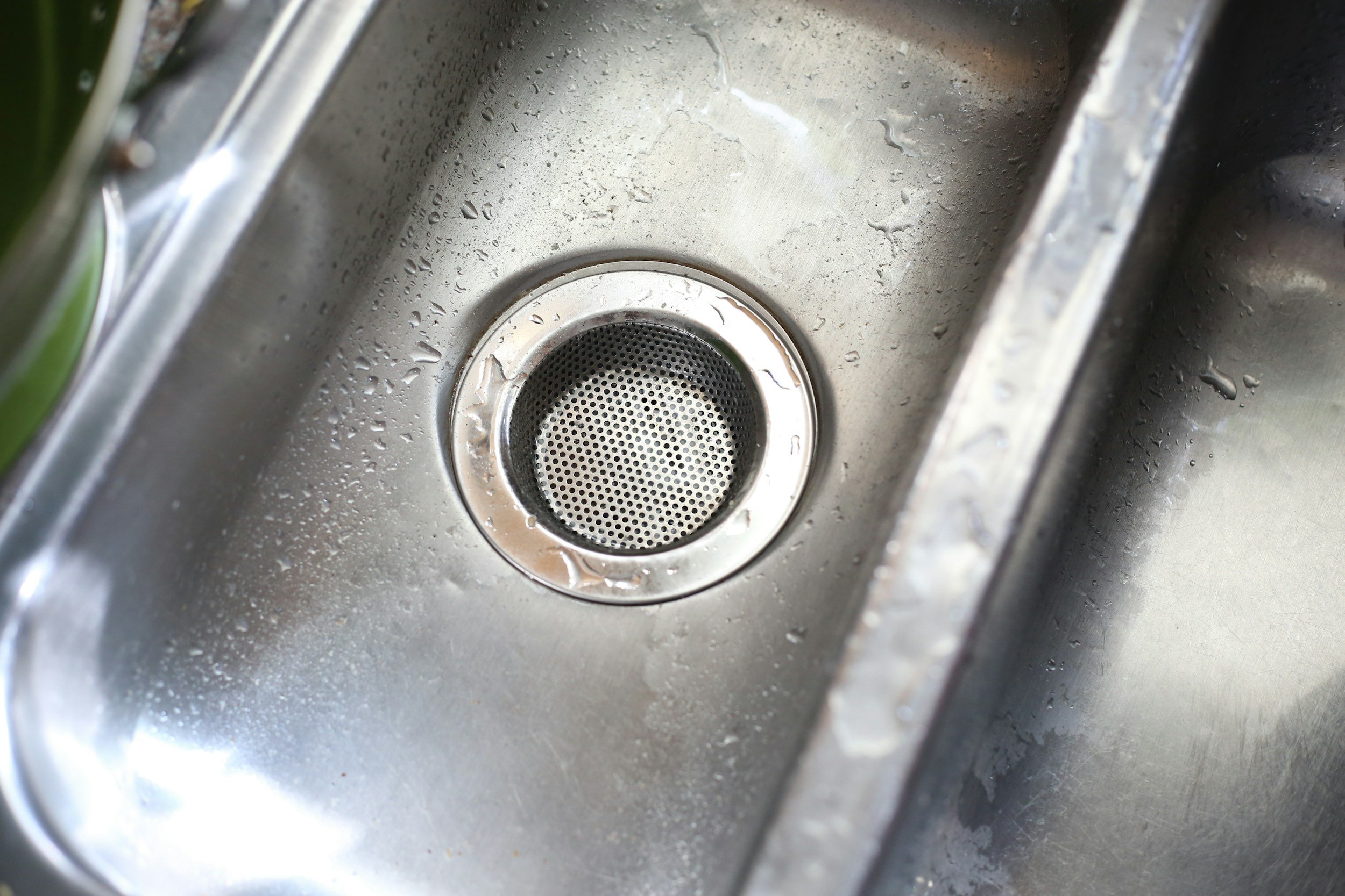 Close-up view of a stainless steel kitchen sink with water droplets, showing the drain with a mesh strainer in the center.