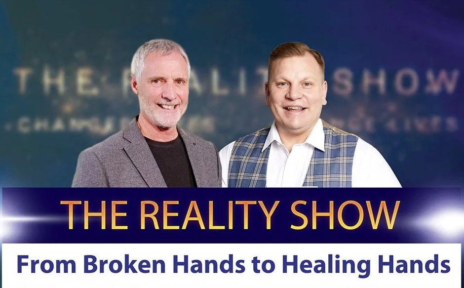 Two men standing in front of a blurred background with the text "THE REALITY SHOW." One man has gray hair and a beard, wearing a blazer, and the other has short red hair, wearing a white shirt and plaid vest. The image includes a banner at the bottom with the text "THE REALITY SHOW" and "From Broken Hands to Healing Hands."