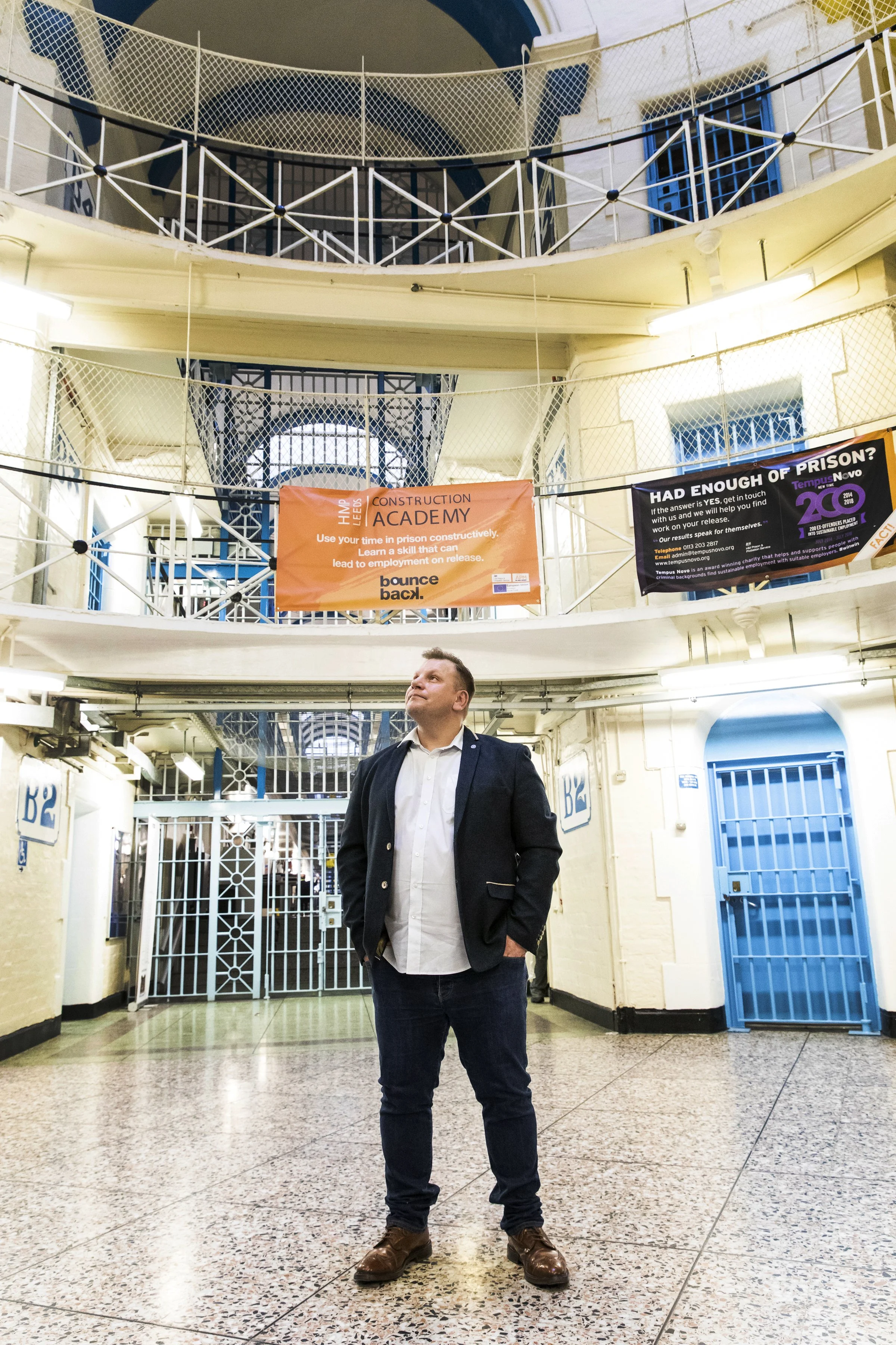 A man in a suit jacket and jeans standing in a prison corridor with blue barred gates and a balcony above. There are signs for a construction academy and prison information.