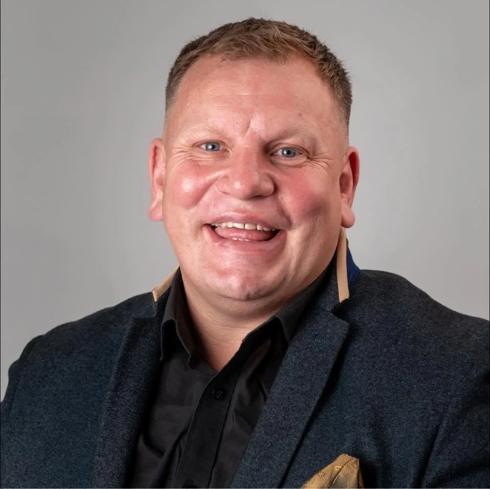 A smiling man with short, light brown hair, wearing a dark suit jacket and black shirt, posing against a light gray background.