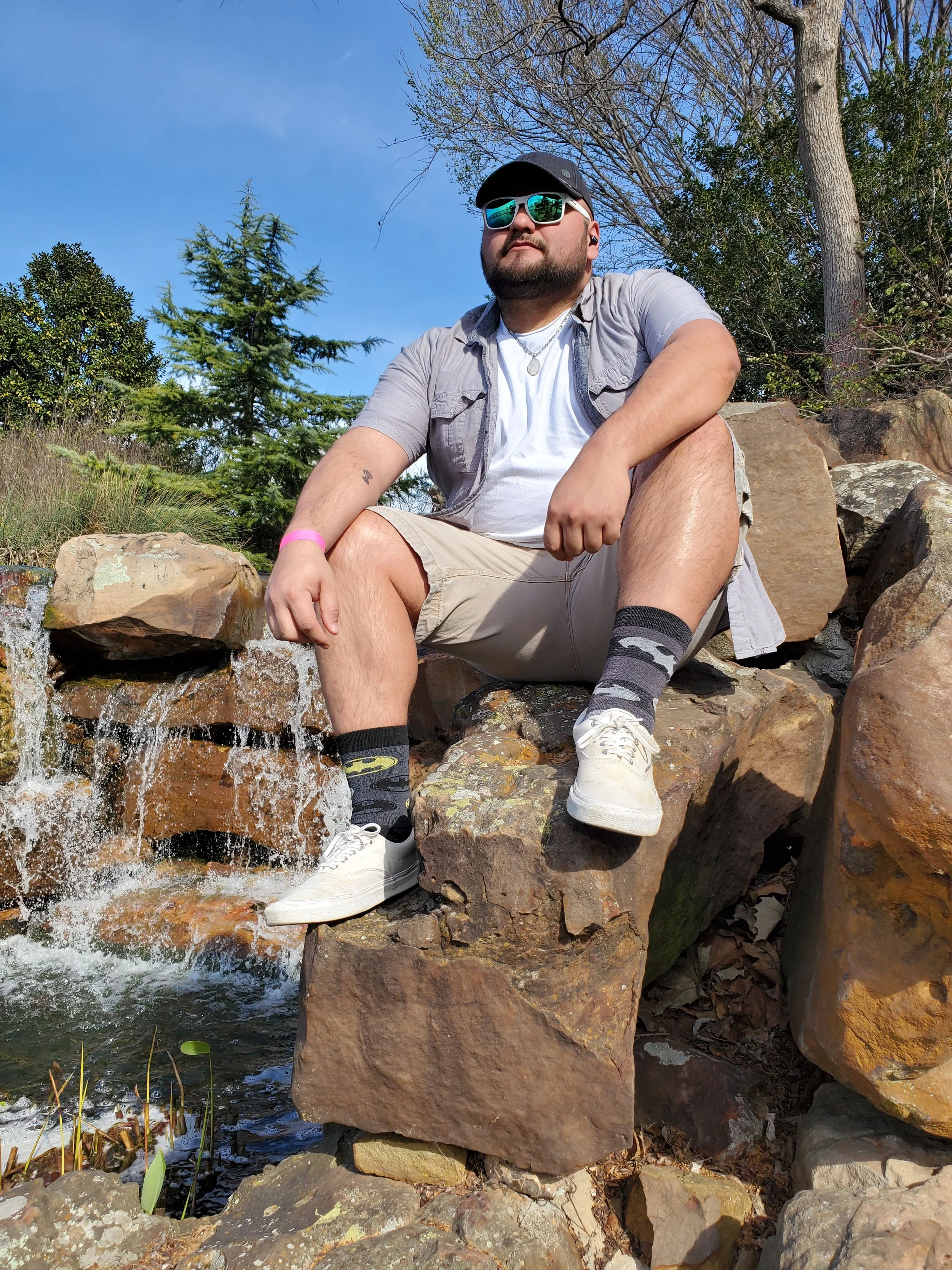 A man sitting on rocks by a small waterfall outdoors, wearing sunglasses, a cap, a light-colored shirt, shorts, and sneakers, with trees and a clear blue sky in the background.