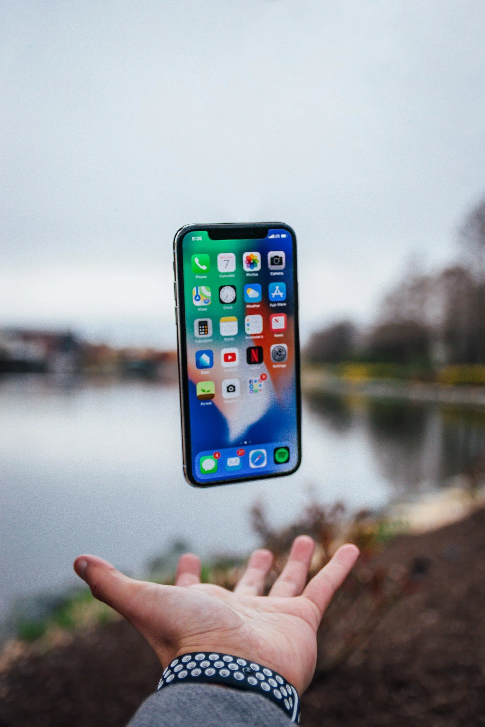 A person wearing a gray long-sleeve shirt and a black wristband with white dots is tossing an iPhone into the air outdoors near a body of water with a blurry cityscape in the background.