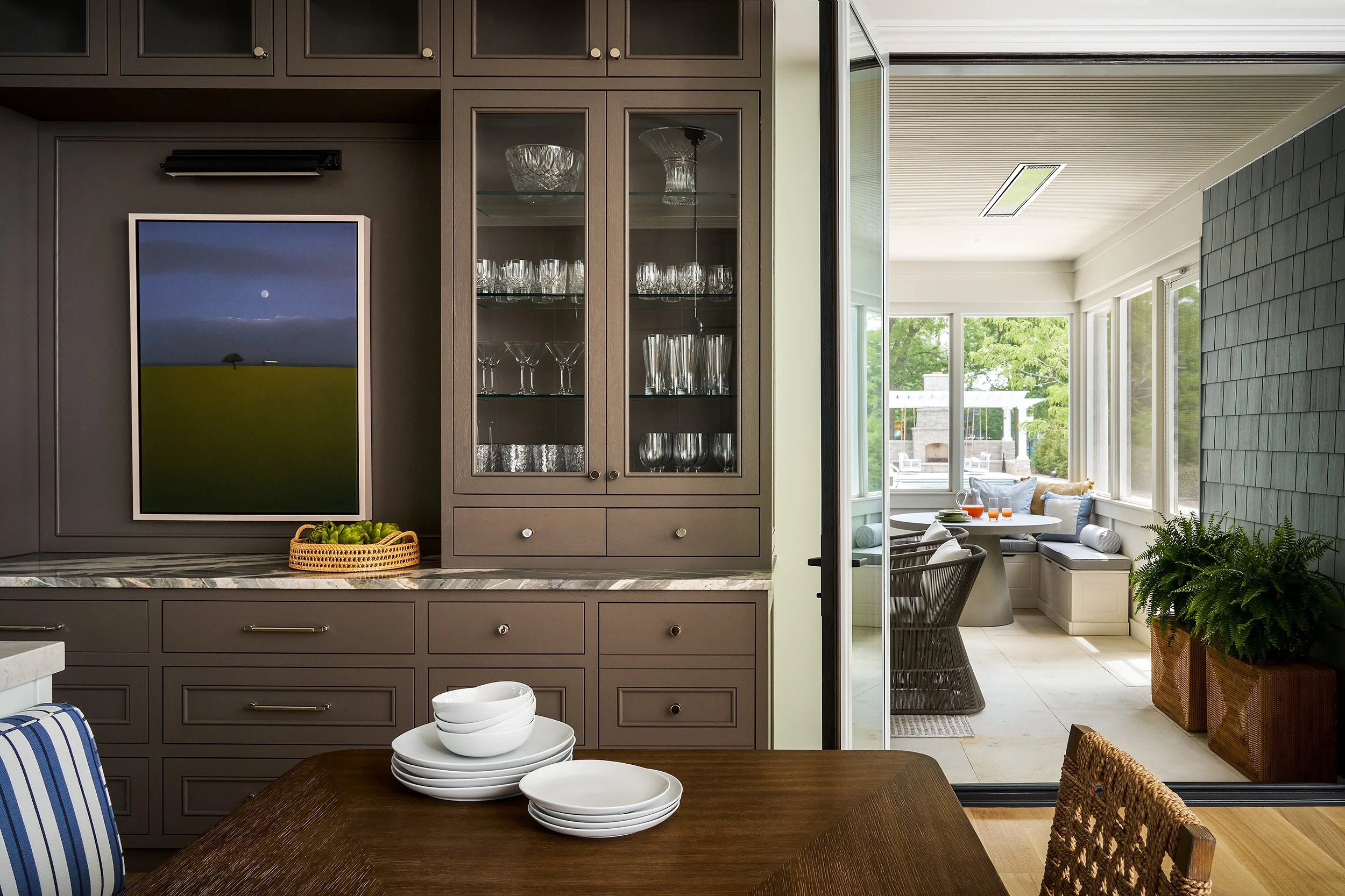 Built-in cabinetry with glass display shelves and a view into a bright sunroom with cozy seating and greenery.