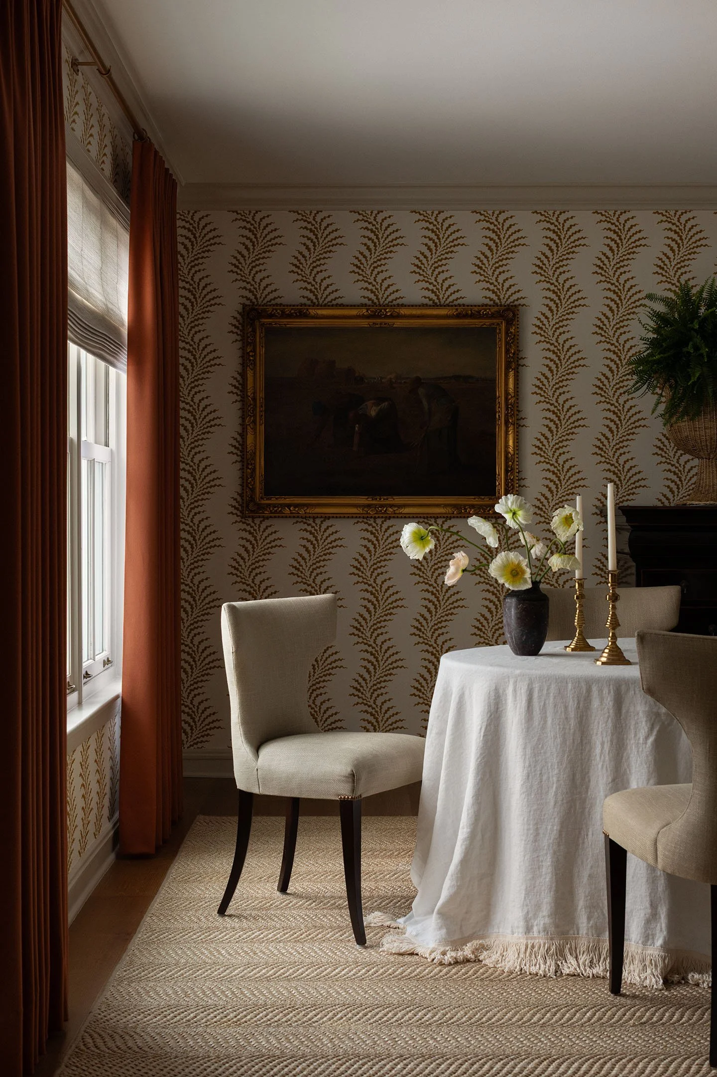 Elegant dining room with patterned wallpaper, framed artwork, rust drapery panels, and round linen-covered table.