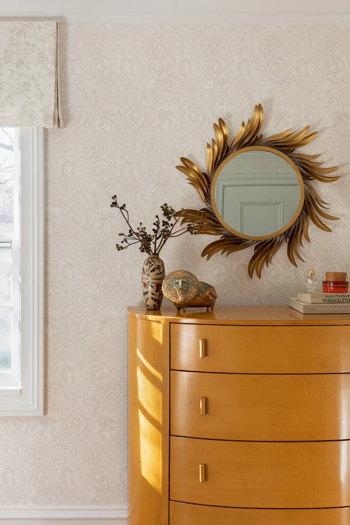 Warm wood dresser styled with decorative vase and gold sunburst mirror against neutral wallpaper.