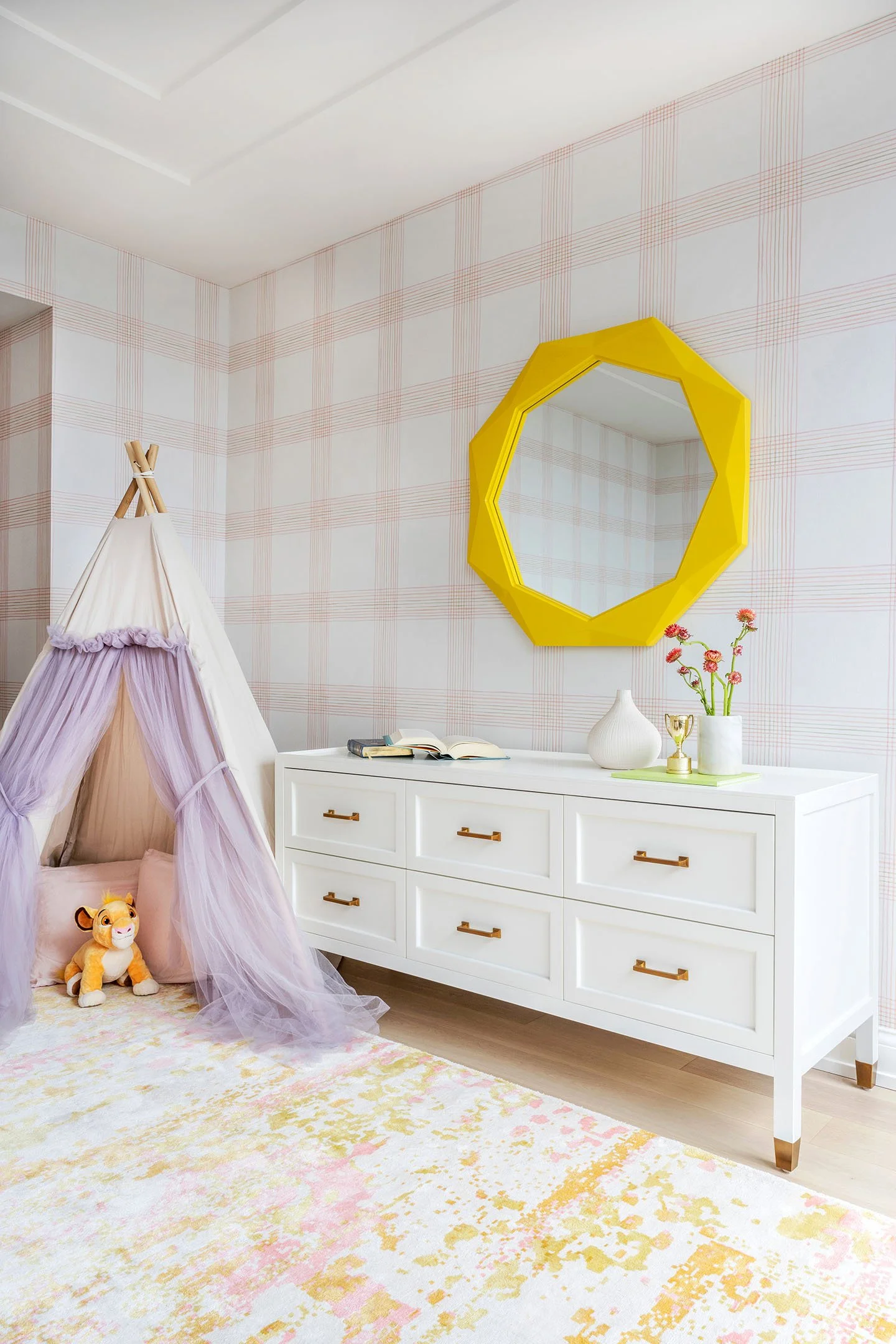Girls bedroom with white six-drawer dresser, geometric yellow mirror, plaid wallpaper, and soft pastel area rug.