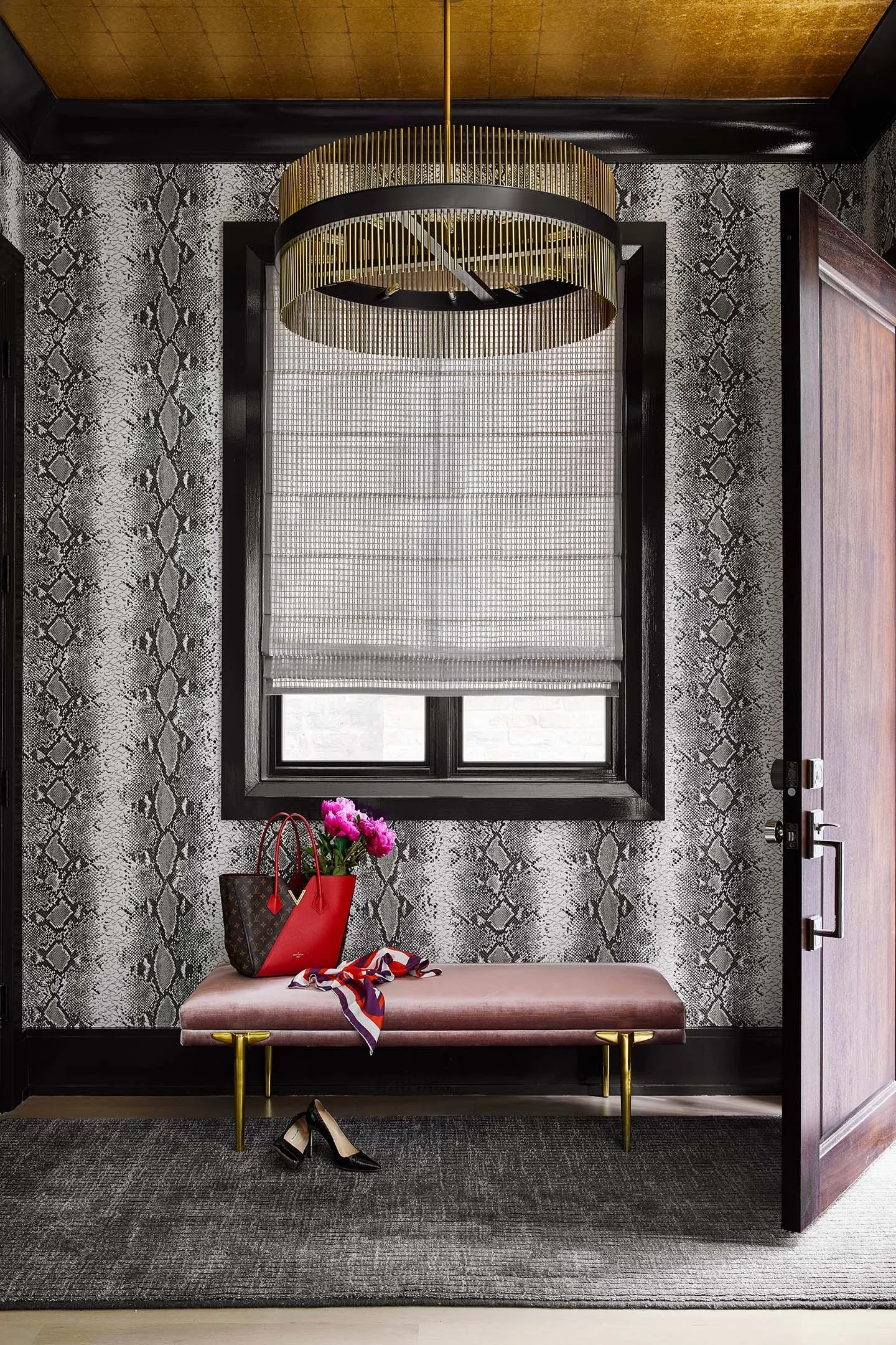 Luxury entryway with upholstered bench, gold chandelier, patterned wallpaper, and designer handbag styling.