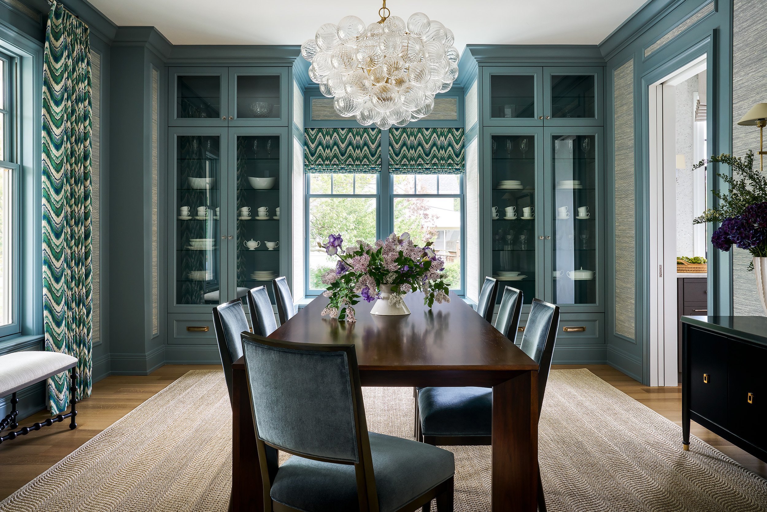 Dining room with teal paneled walls, patterned drapery, a wooden table, and a decorative glass chandelier.