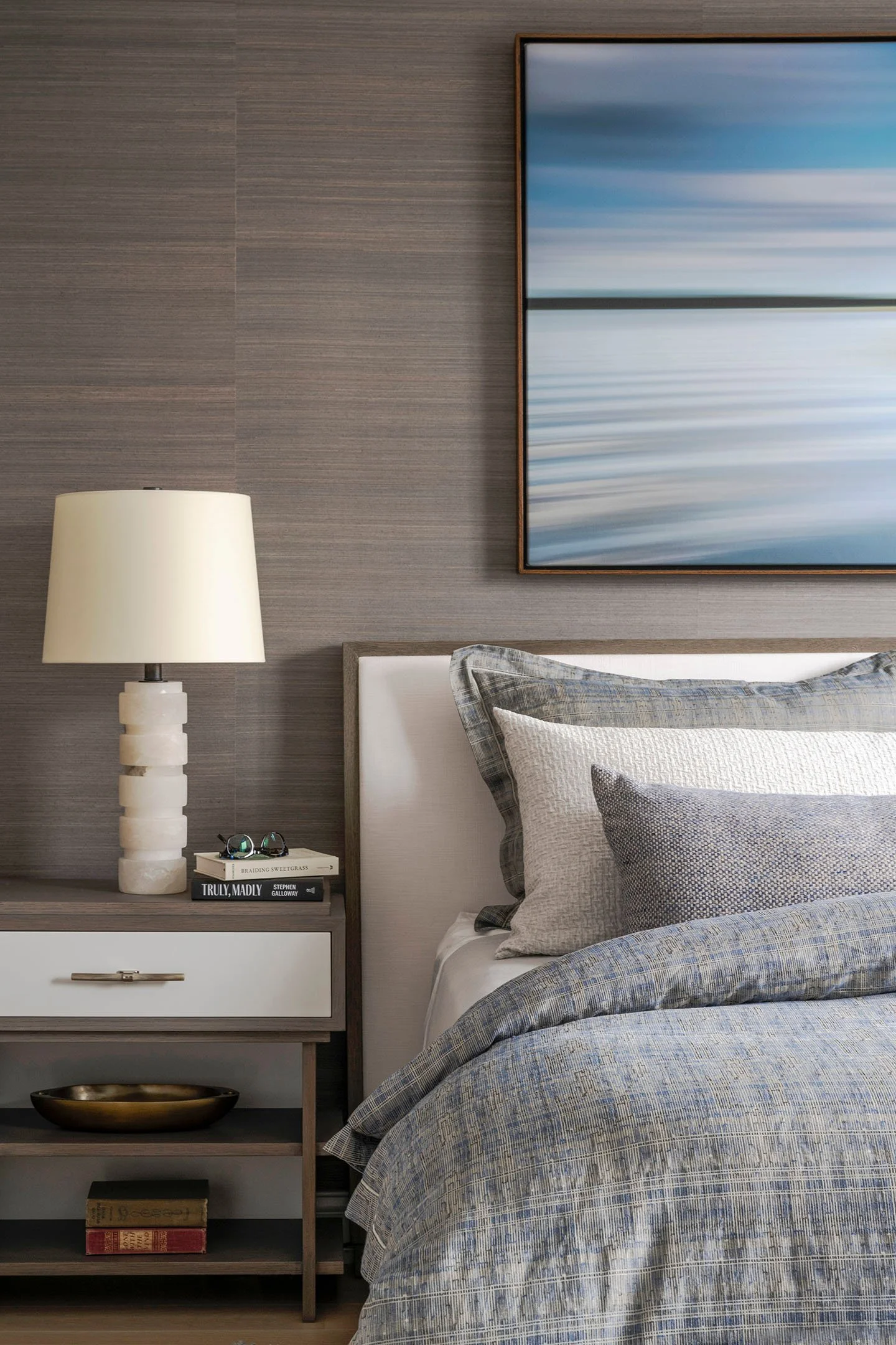 Modern primary bedroom detail with upholstered headboard, layered bedding, wood nightstand, sculptural table lamp, and abstract artwork.