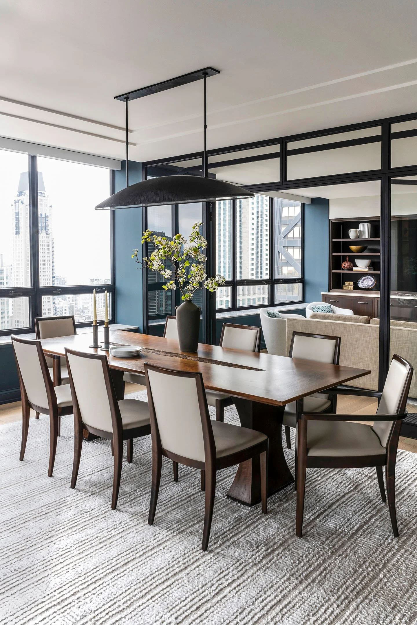 Luxury dining room featuring a solid wood table, cream upholstered chairs, oversized black pendant light, and panoramic city views.