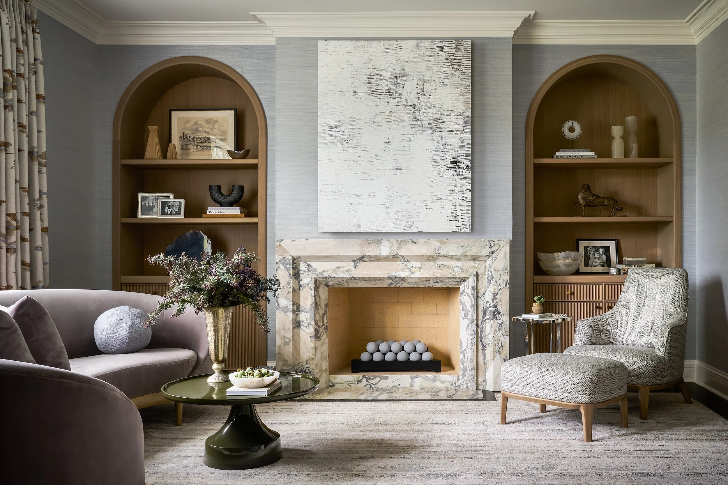 A refined living room featuring a statement marble fireplace with layered stone surround, arched wood built-ins styled with curated decor, and a curved blush-toned sofa paired with a sculptural glass coffee table.